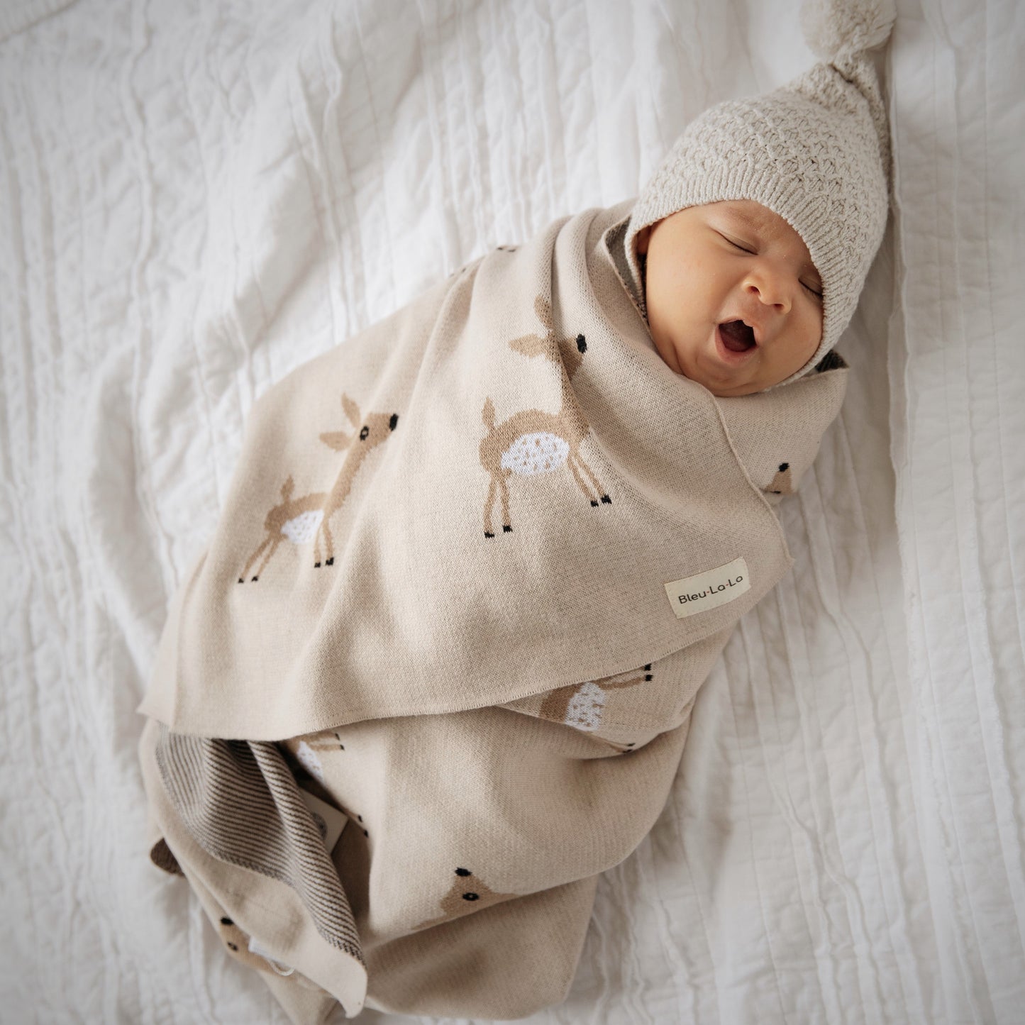 Newborn baby wrapped in a beige blanket with animal patterns, wearing a matching hat, on a white textured surface.