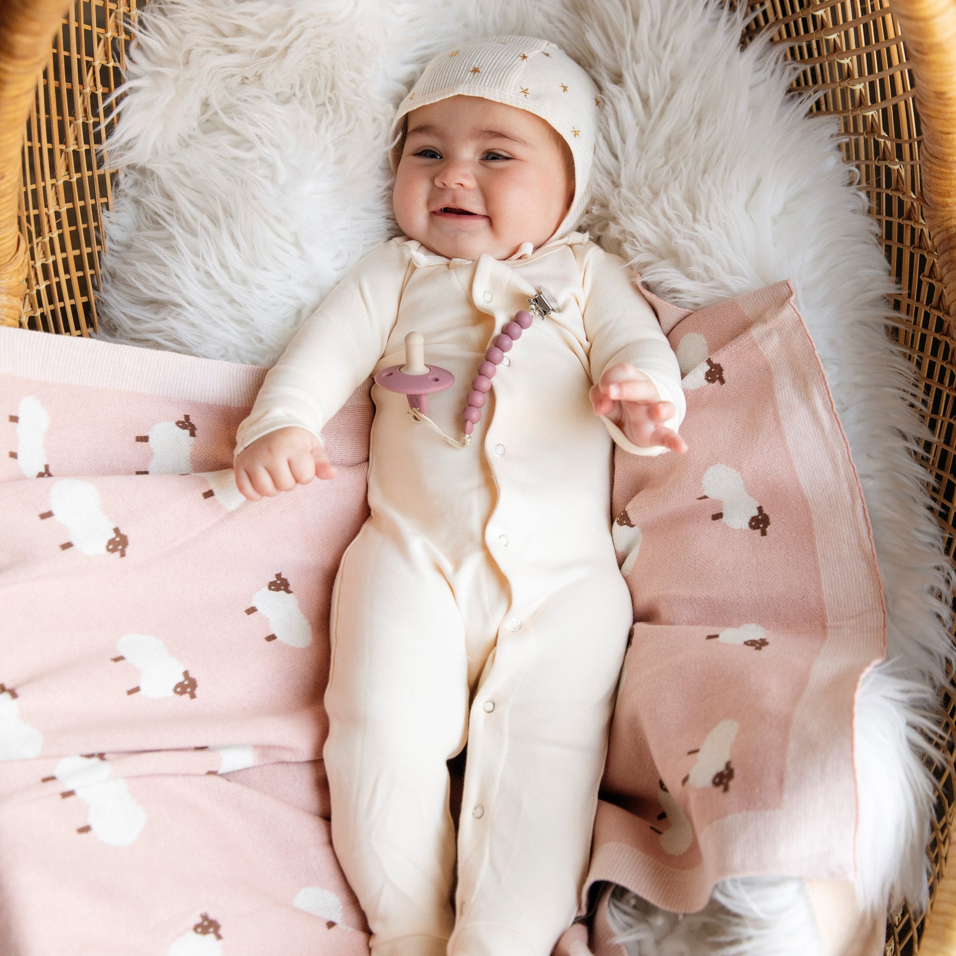 Baby in a cream-colored outfit with a hood, sitting on a pink blanket with sheep patterns, from 'Bleu La La',  on a fluffy white surface.