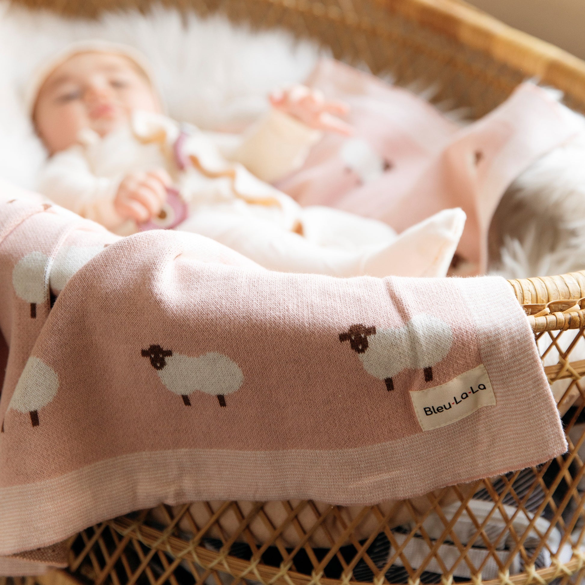 Baby lying in a crib with a pink blanket featuring sheep designs from 'Bleu La La'.
