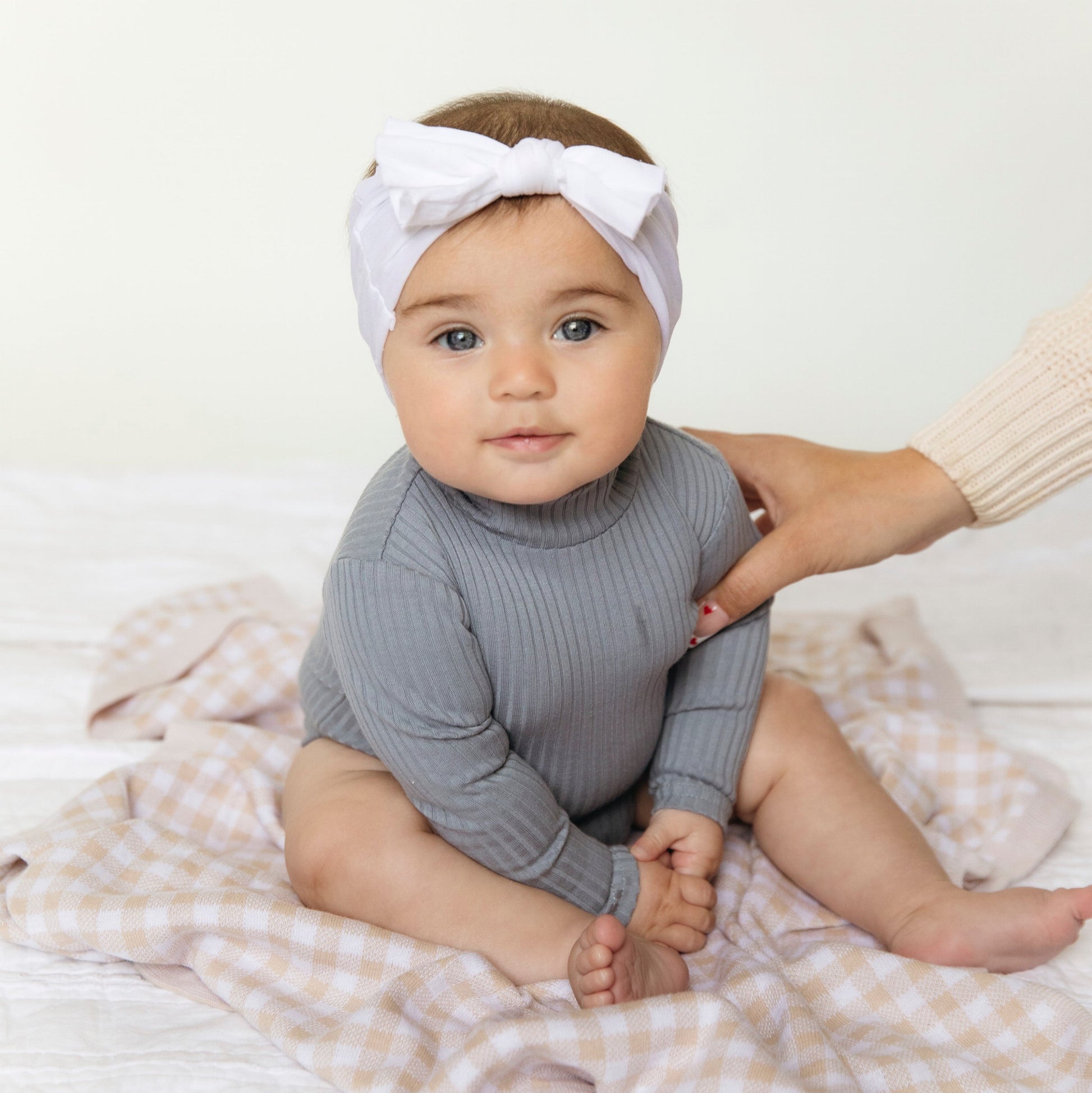 Baby wearing a gray ribbed outfit and white headband with a bow, sitting on a checkered blanket.