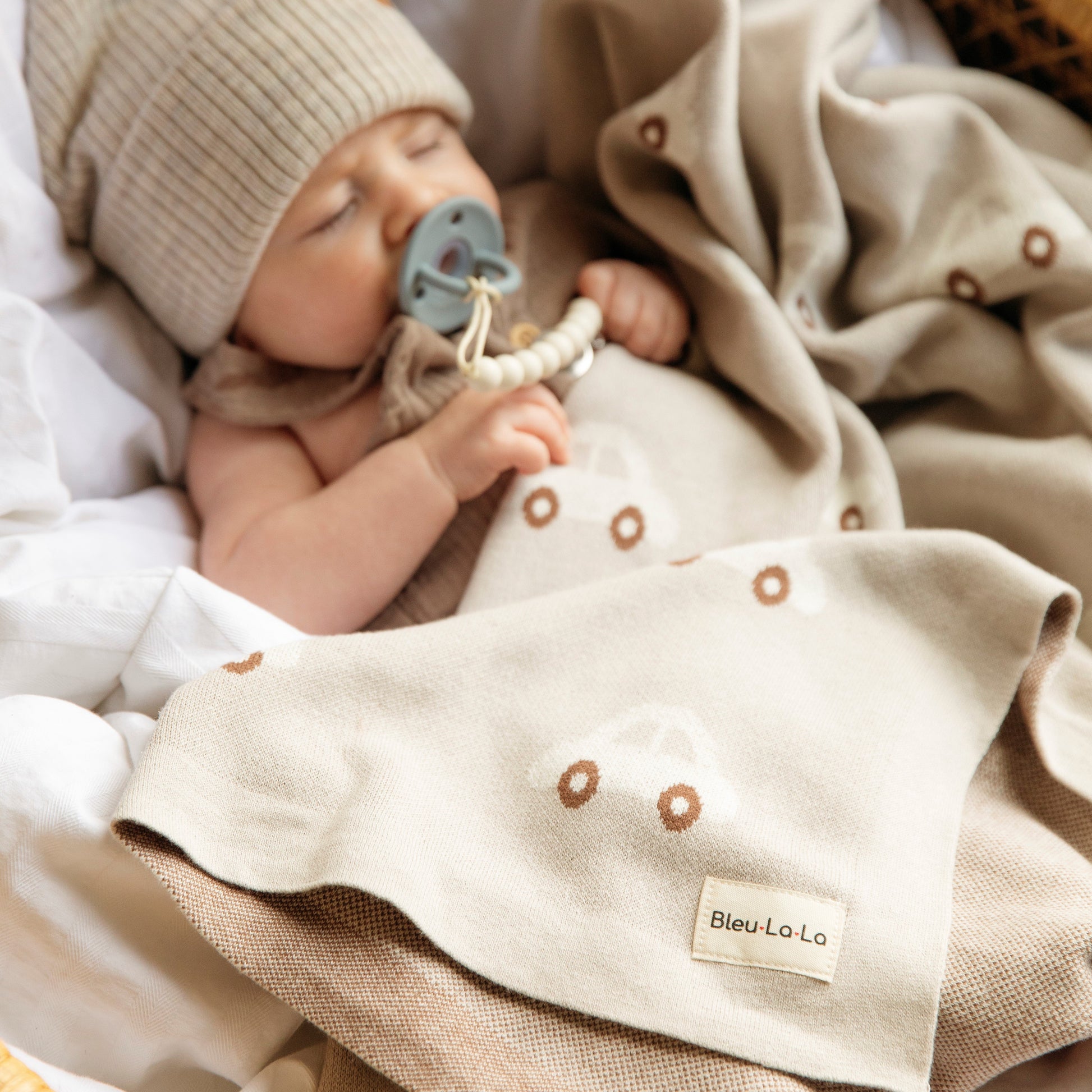 Baby wrapped in a blanket with a 'Bleu-La-La' label, wearing a beige knit hat and holding a blue pacifier.