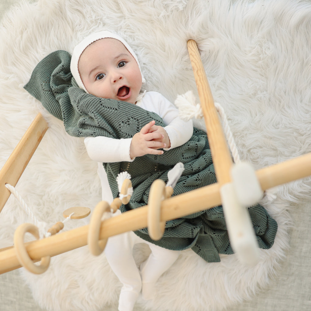 Baby in a green knitted outfit and white hat holding a green heart Bleu-la-la  blanket with a fluffy white background