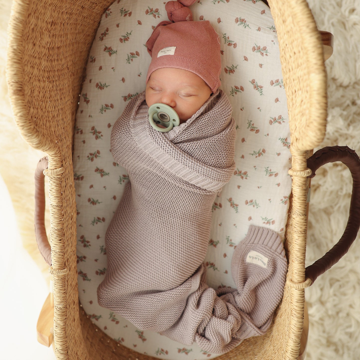 Newborn baby swaddled in a pink Blue-La-La blanket with a pacifier, wearing a matching pink hat, in a wicker basket with a floral patterned sheet.