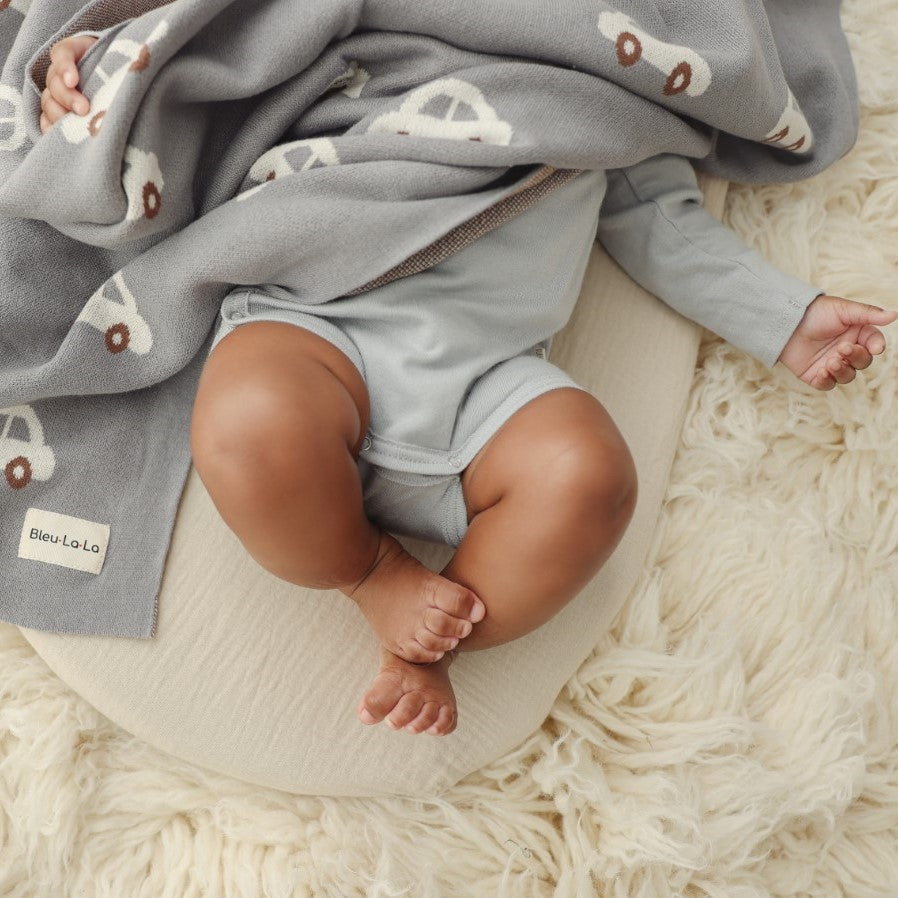 Baby in light gray outfit sitting on a cushion with a blanket featuring car patterns.