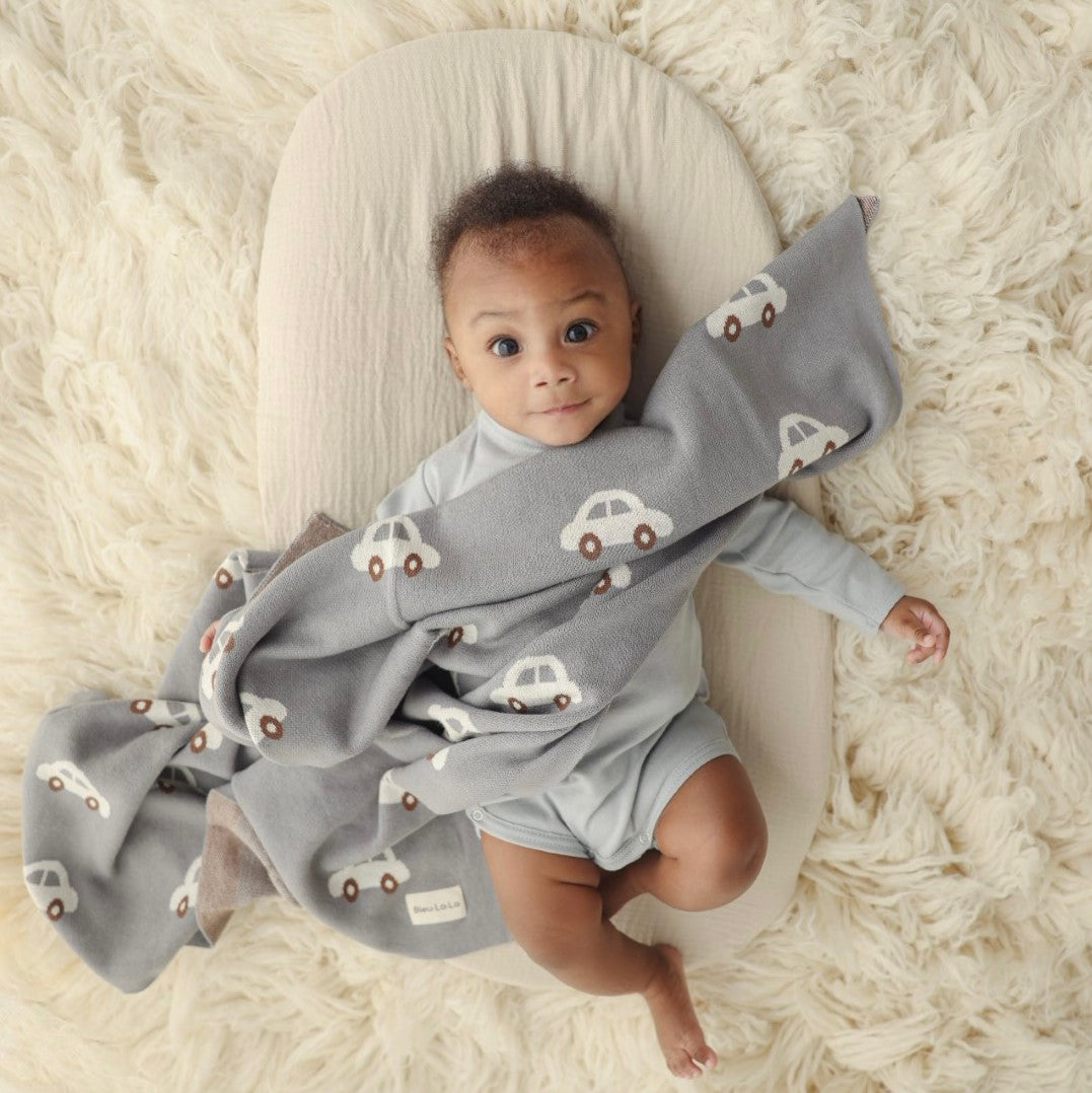 Baby wrapped in a gray blanket with car patterns, lying on a white cushion on a soft white background