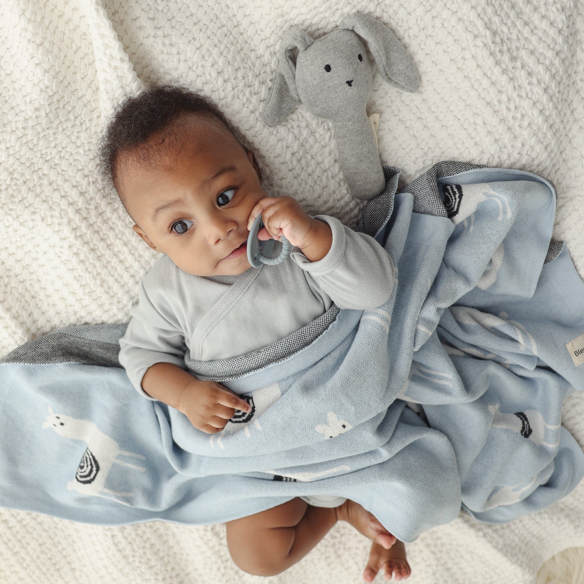 Baby wrapped in a blue blanket with a gray toy, lying on a soft surface