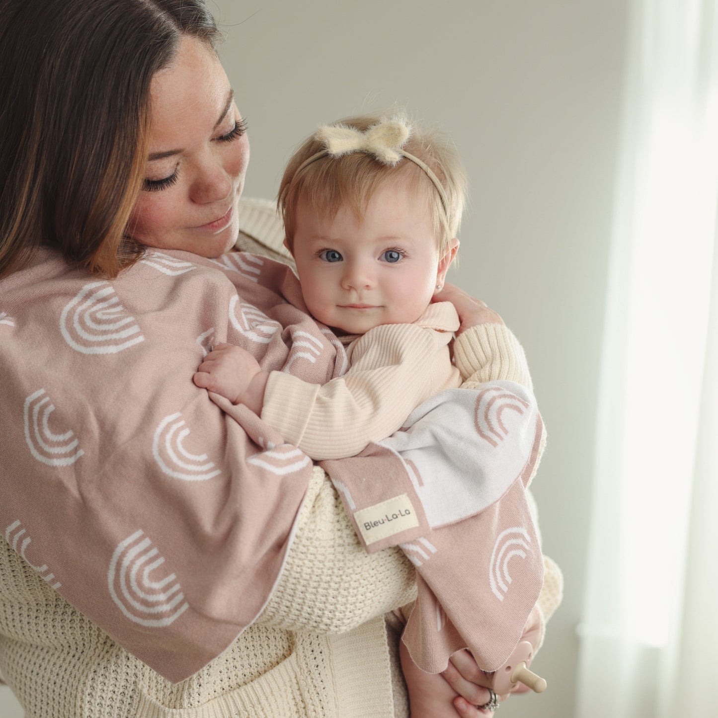 Woman holding a baby wrapped in a pink blanket with white rainbow patterns, indoors.