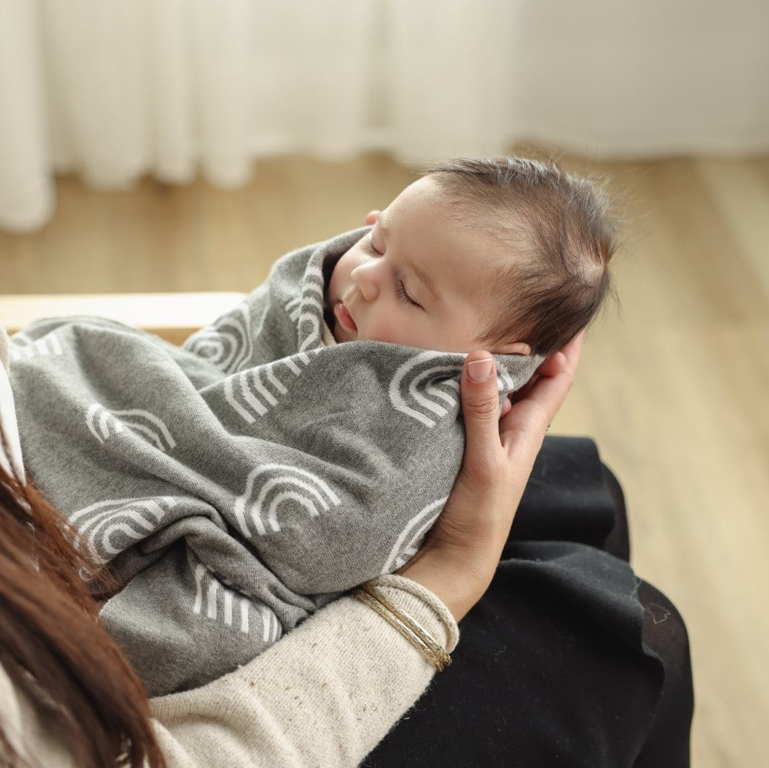 Newborn baby wrapped in a gray blanket with white rainbow patterns, held by an adult in a softly blurred indoor setting.