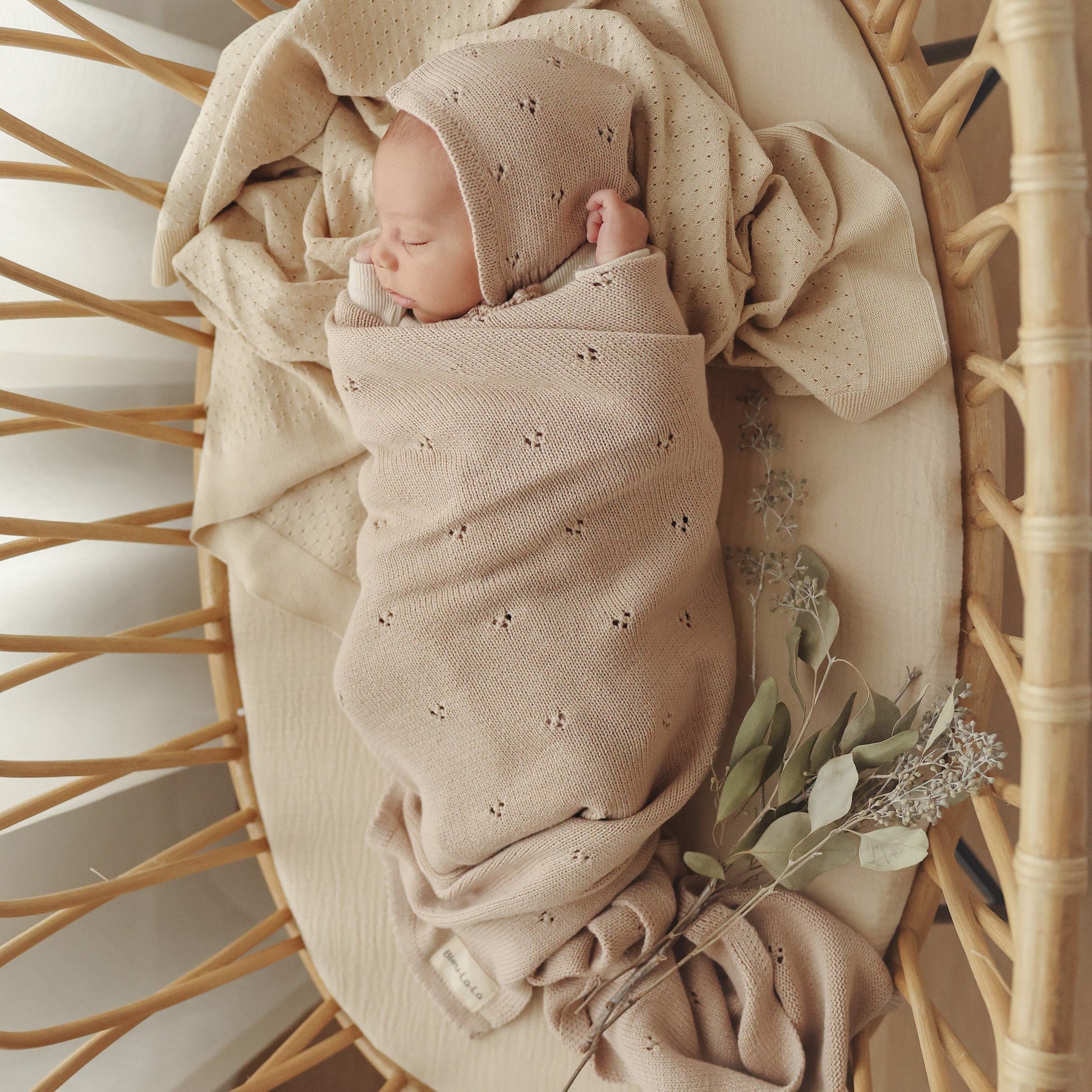 Newborn baby wrapped in a beige swaddle with a matching bonnet, lying on a wooden basket.