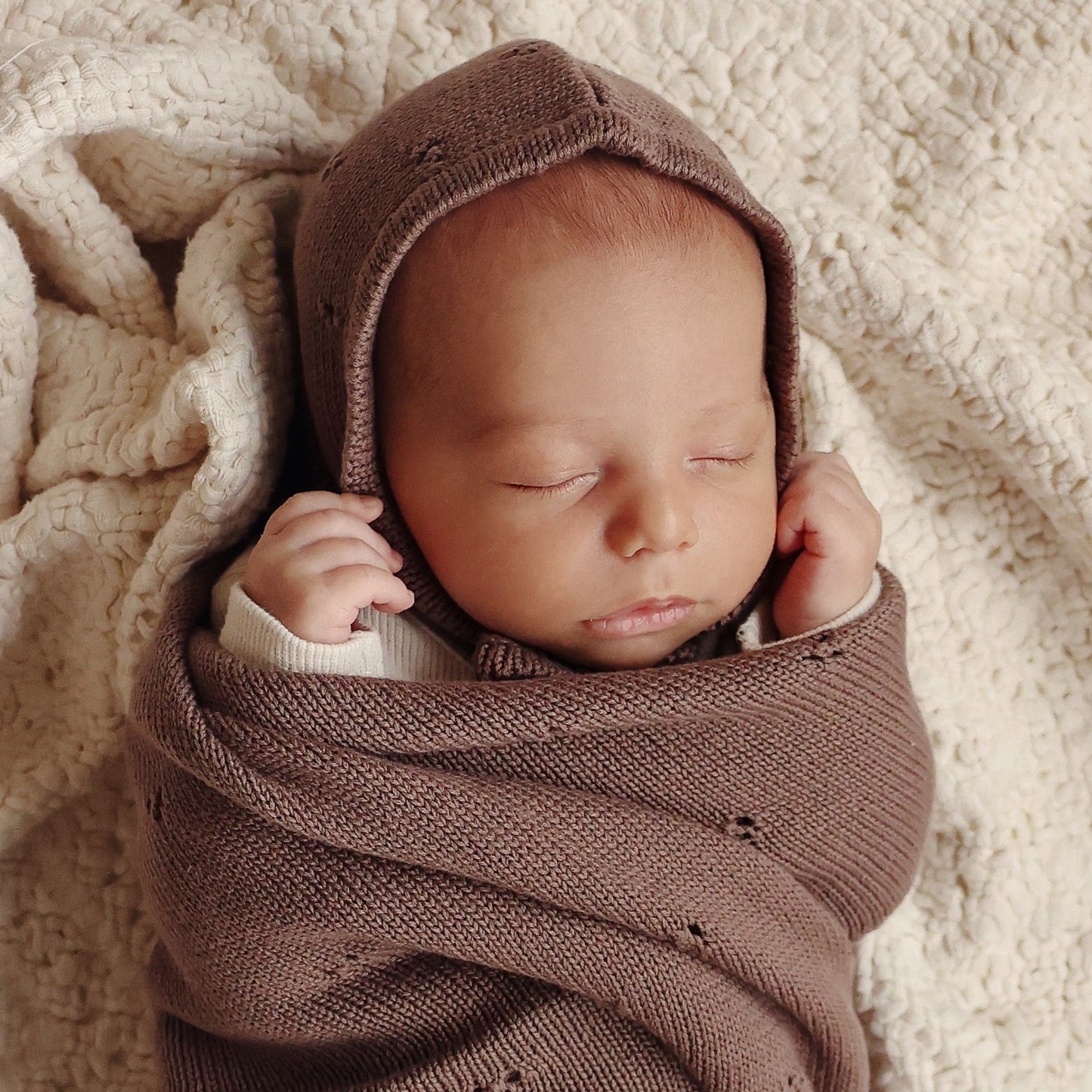 Newborn baby wrapped in a brown knitted blanket and bonnet from on a soft beige surface