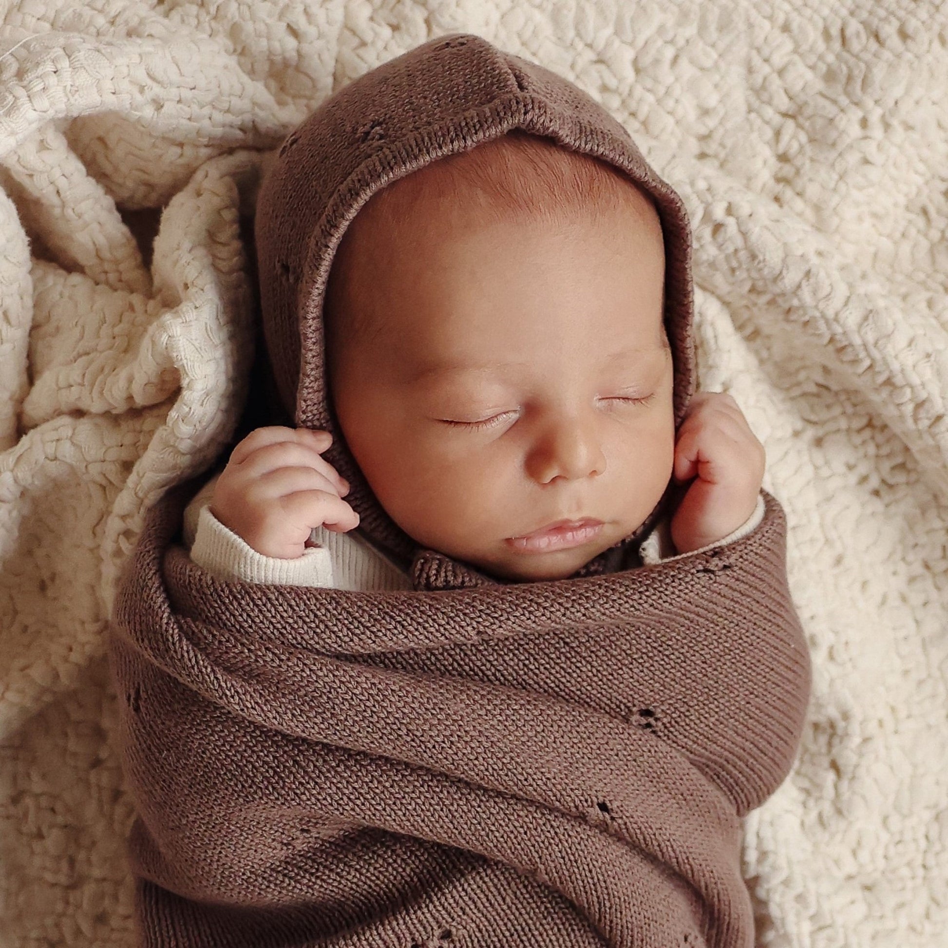 Newborn baby wrapped in a brown knitted blanket and bonnet on a soft beige surface