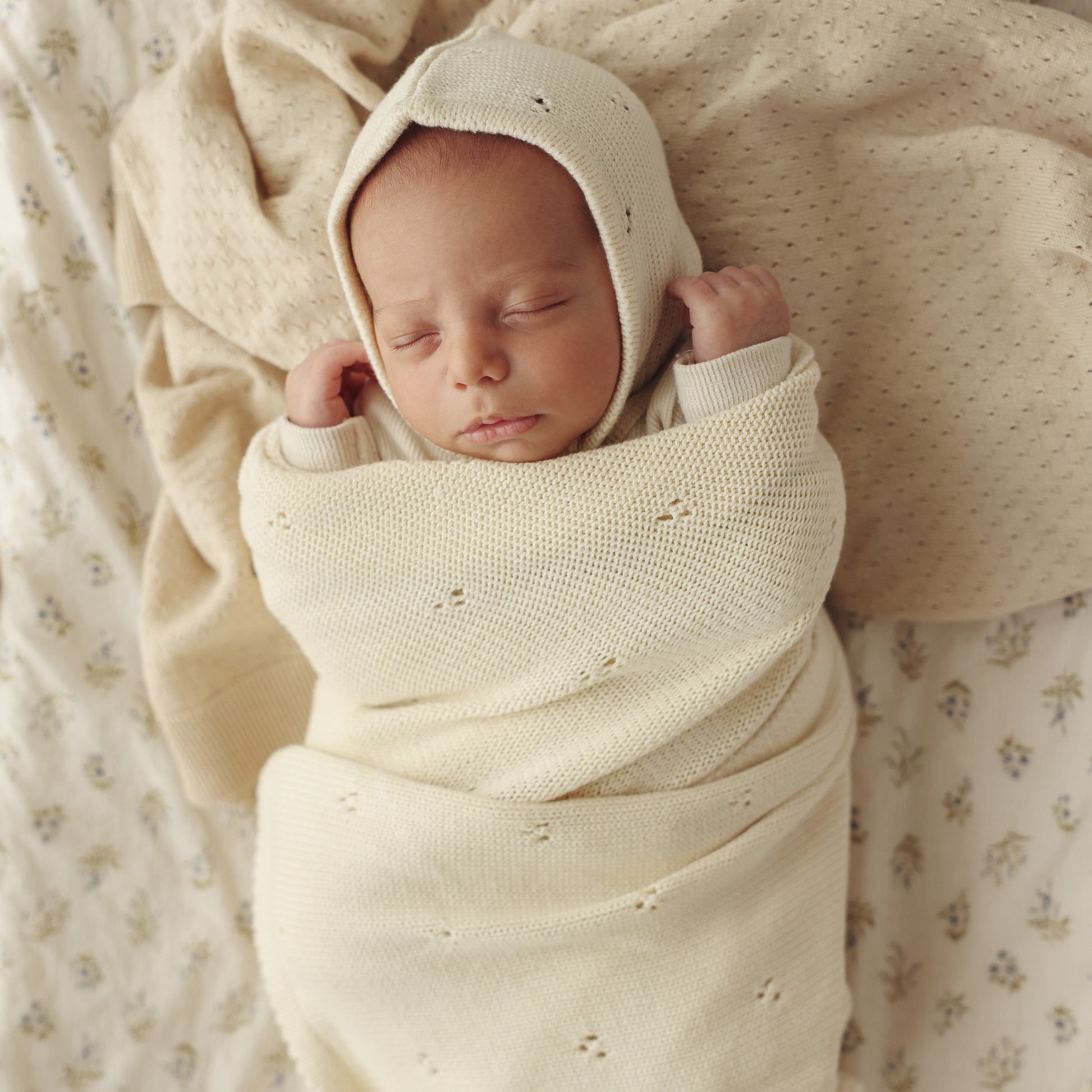 Newborn baby wrapped in a cream-colored swaddle and bonnet on a soft, patterned blanket.