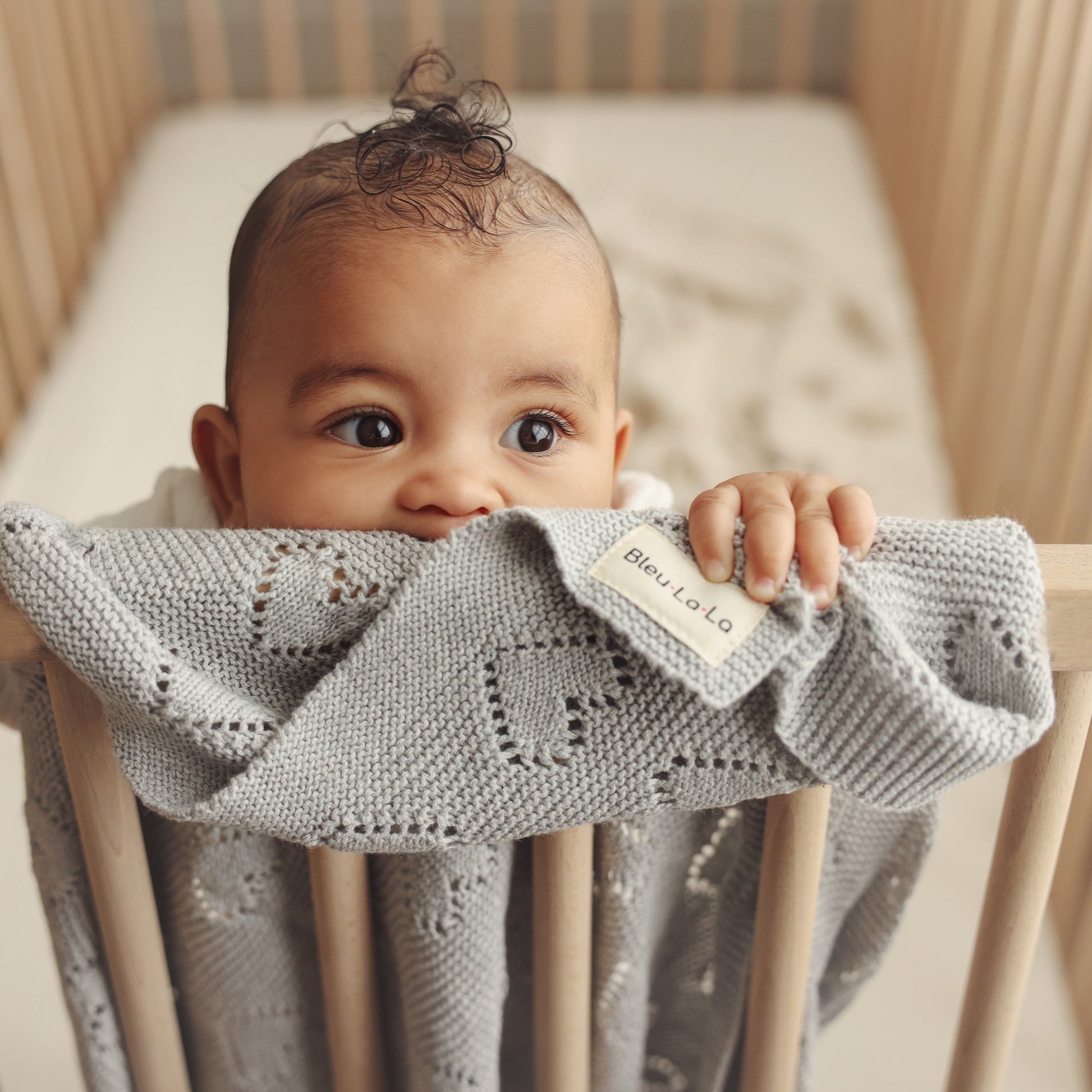 Baby peeking from behind a gray knit Bleu-la-la blanket with a visible brand label in a crib.
