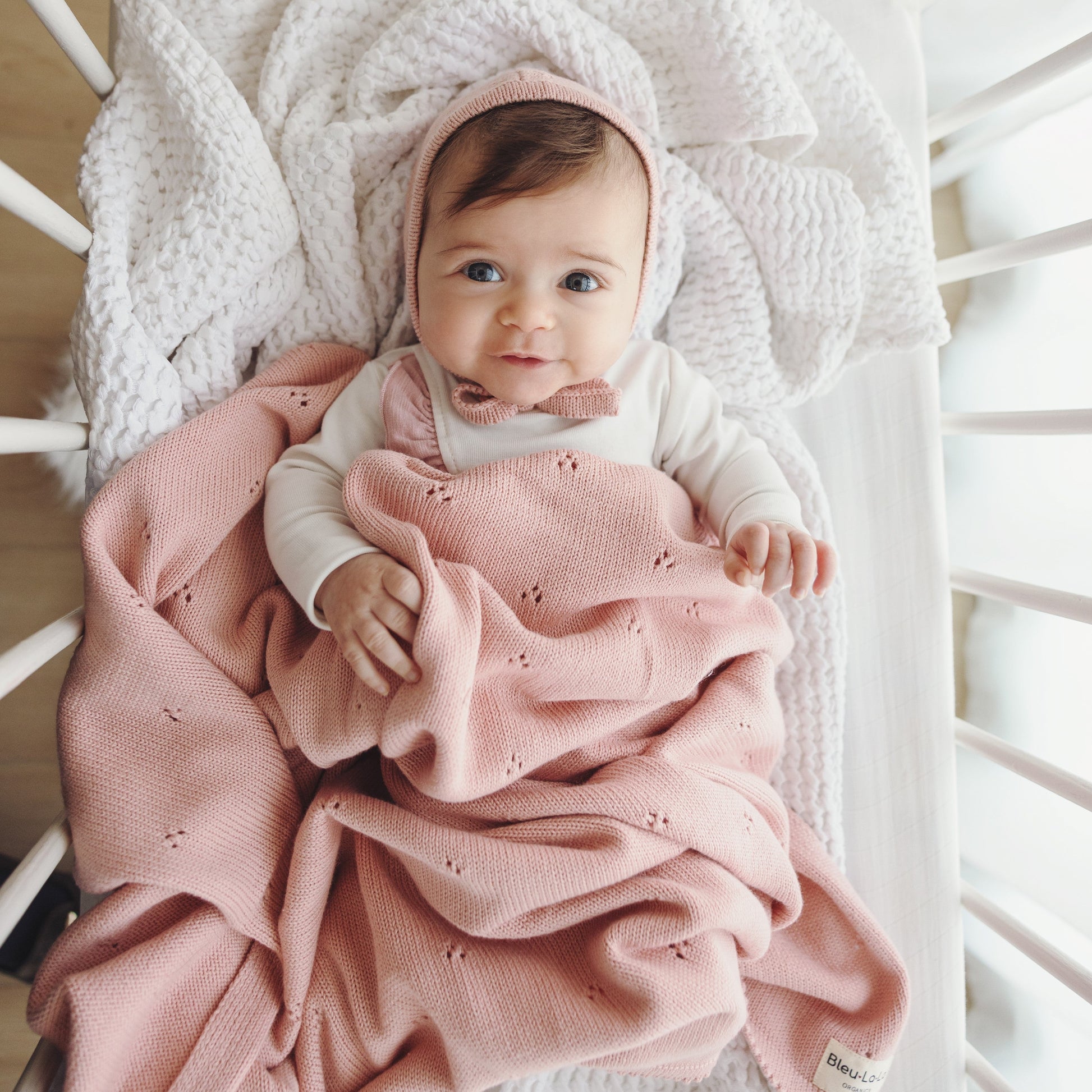 Baby wrapped in a pink blanket and hooded outfit lying in a crib.