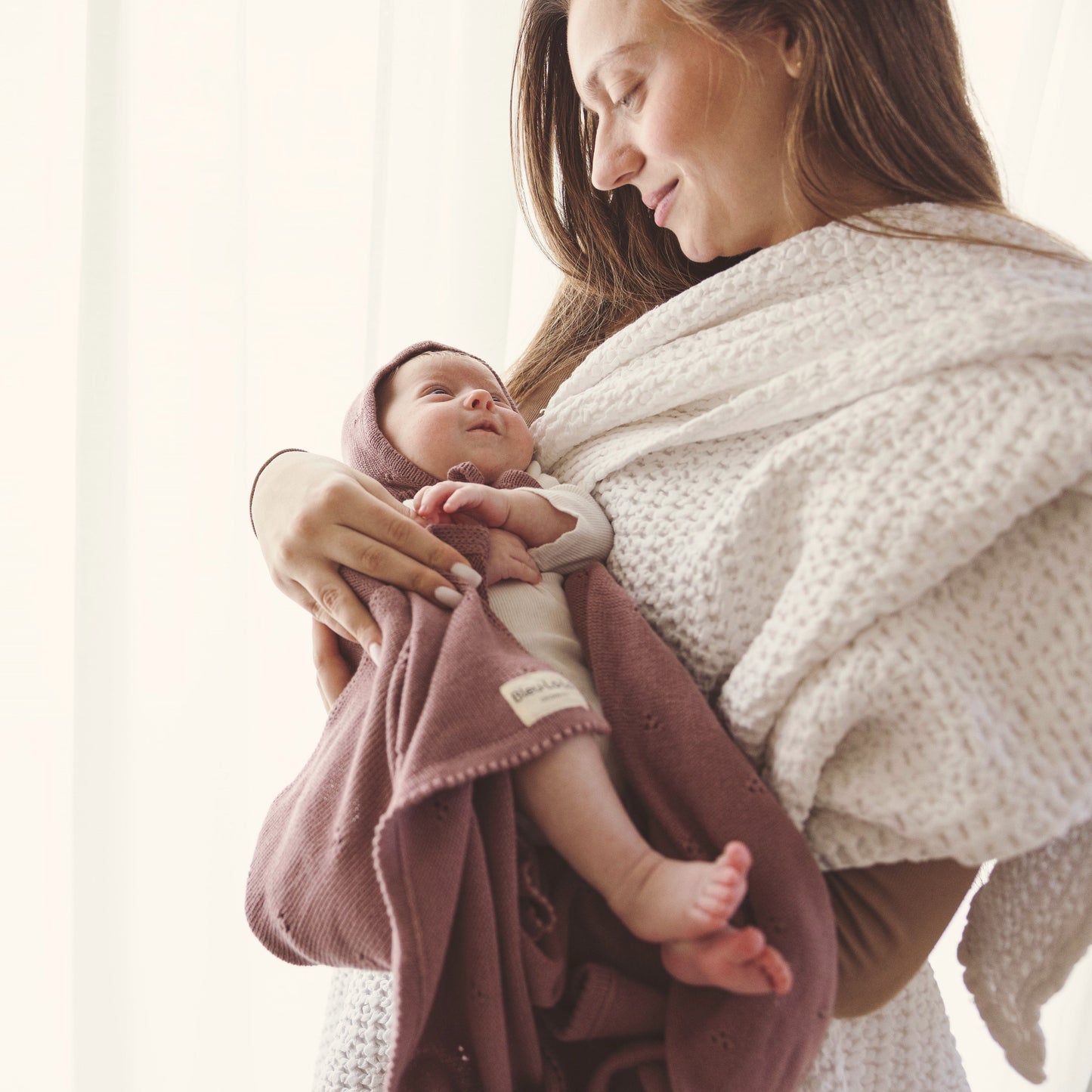 A woman holding a baby wrapped in a pink blanket with a white textured wrap.