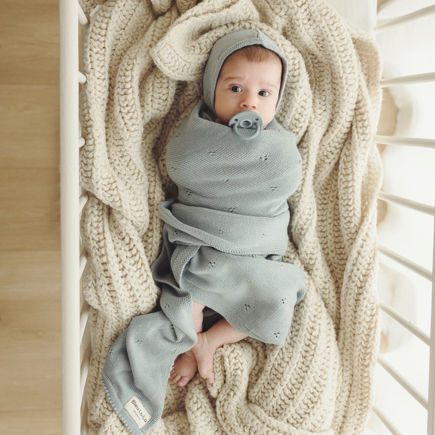 Newborn baby wrapped in a gray swaddle blanket with a pacifier, lying on a beige knitted blanket.
