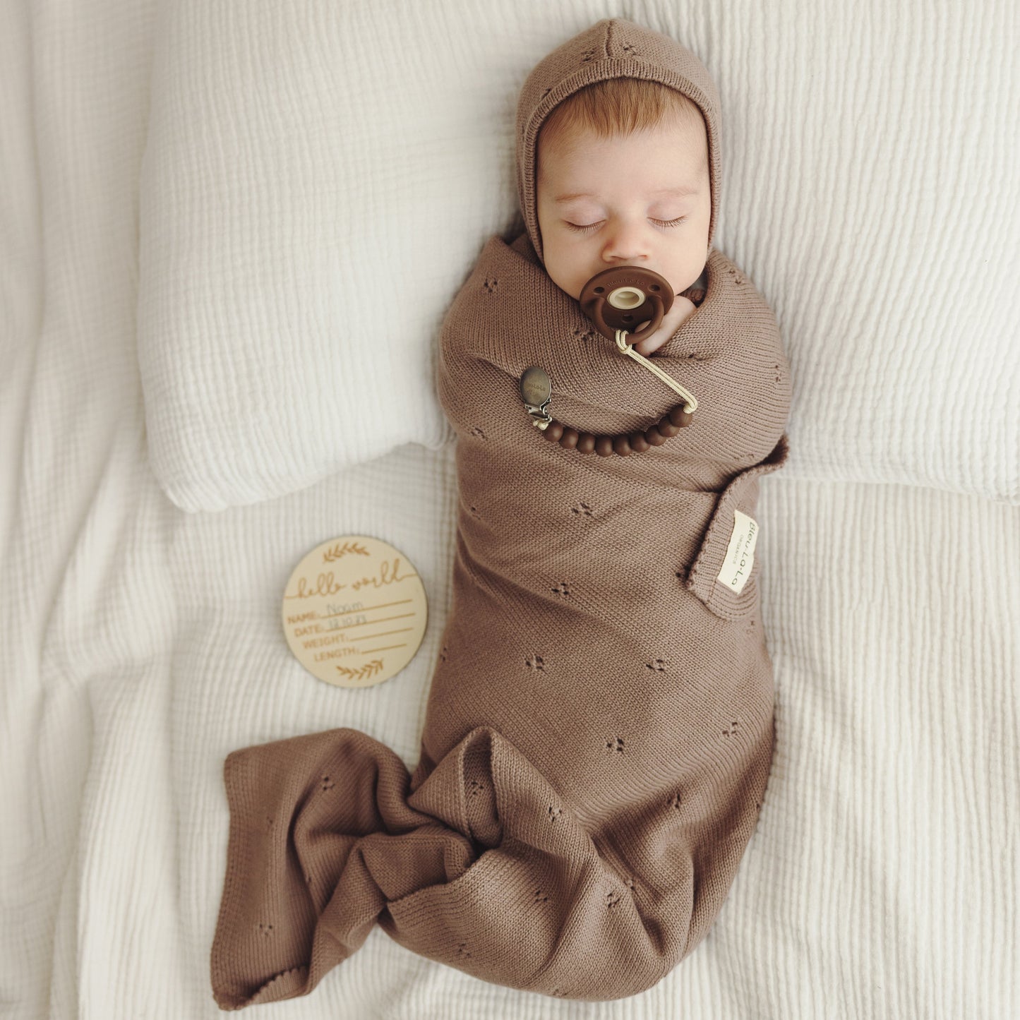 Newborn baby swaddled in brown blanket with pacifier, lying on light-colored surface.