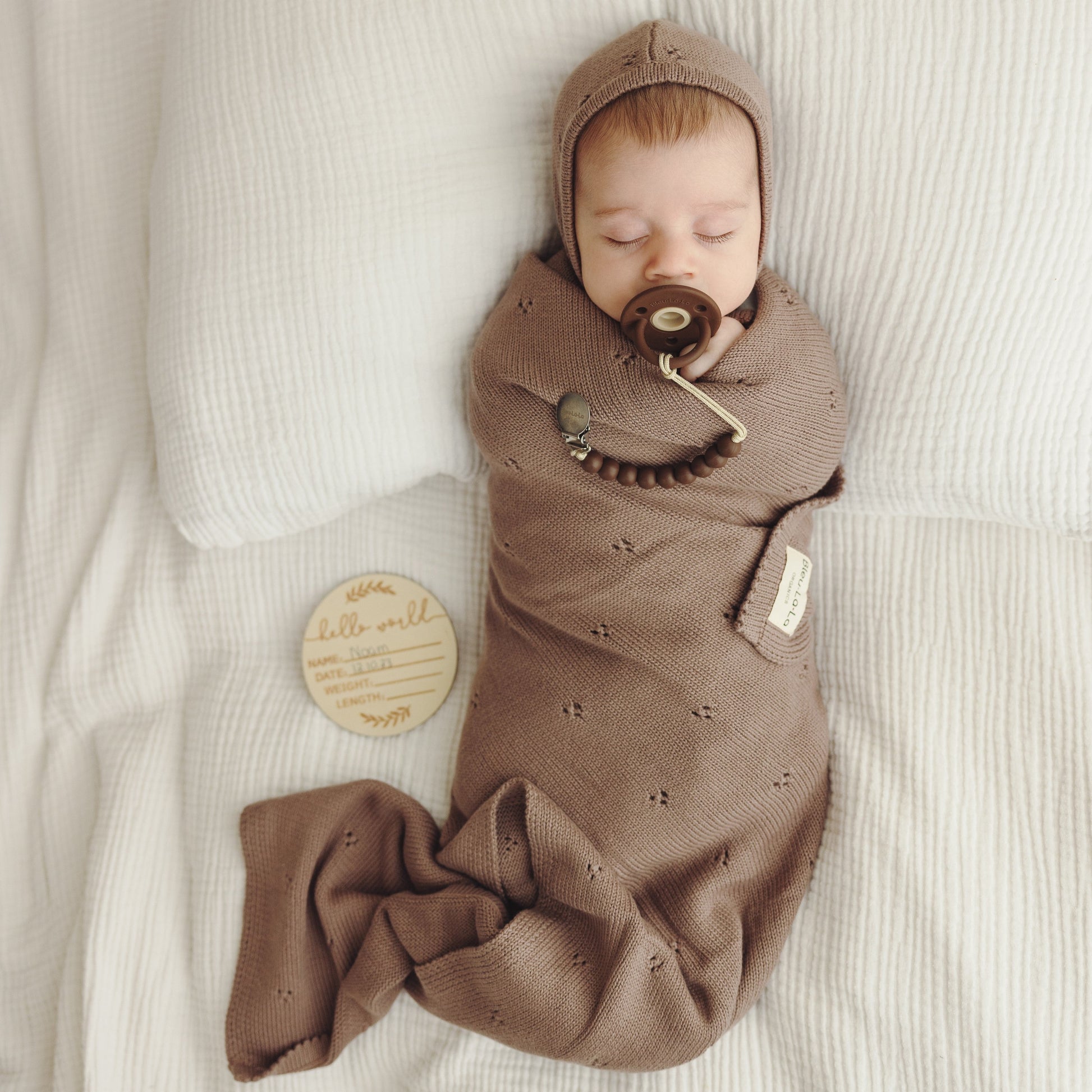 Newborn baby swaddled in brown blanket with pacifier, lying on light-colored surface.