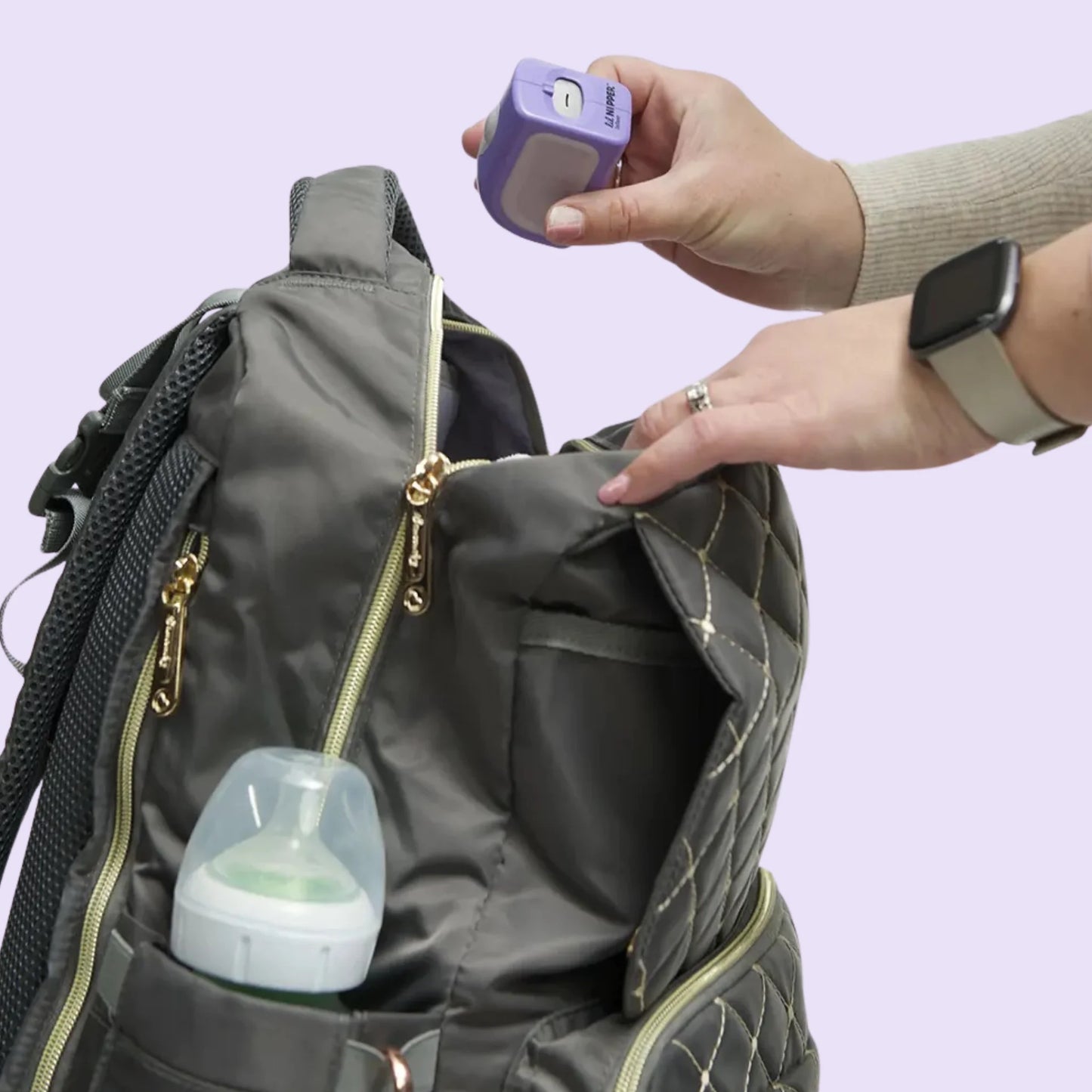 Female placing a purple Lil Nipper device into a gray diaper bag with a bottle inside, on a light purple background.