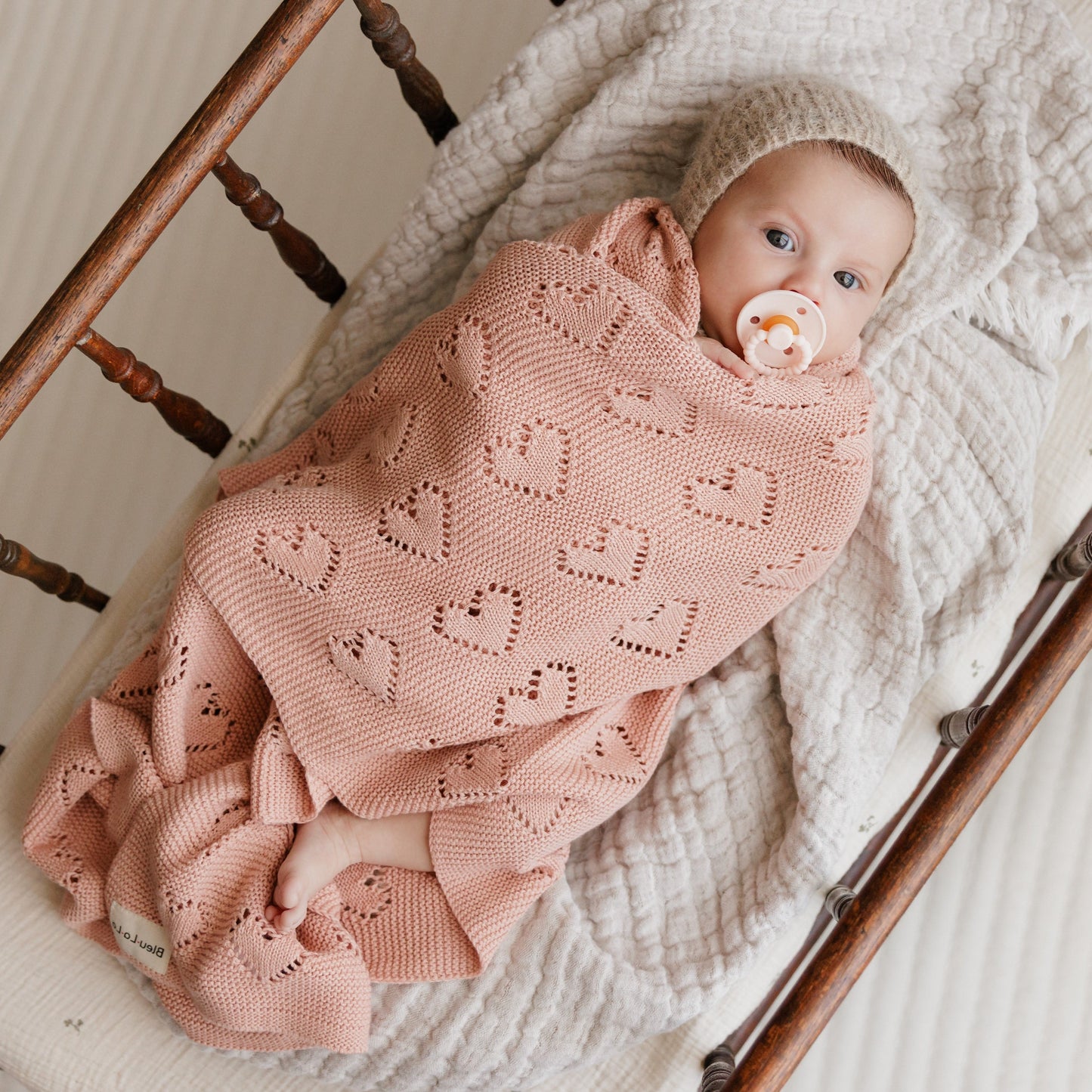 Baby wrapped in a pink knitted Bleu-la-la  blanket with a pacifier, lying on a wooden crib.