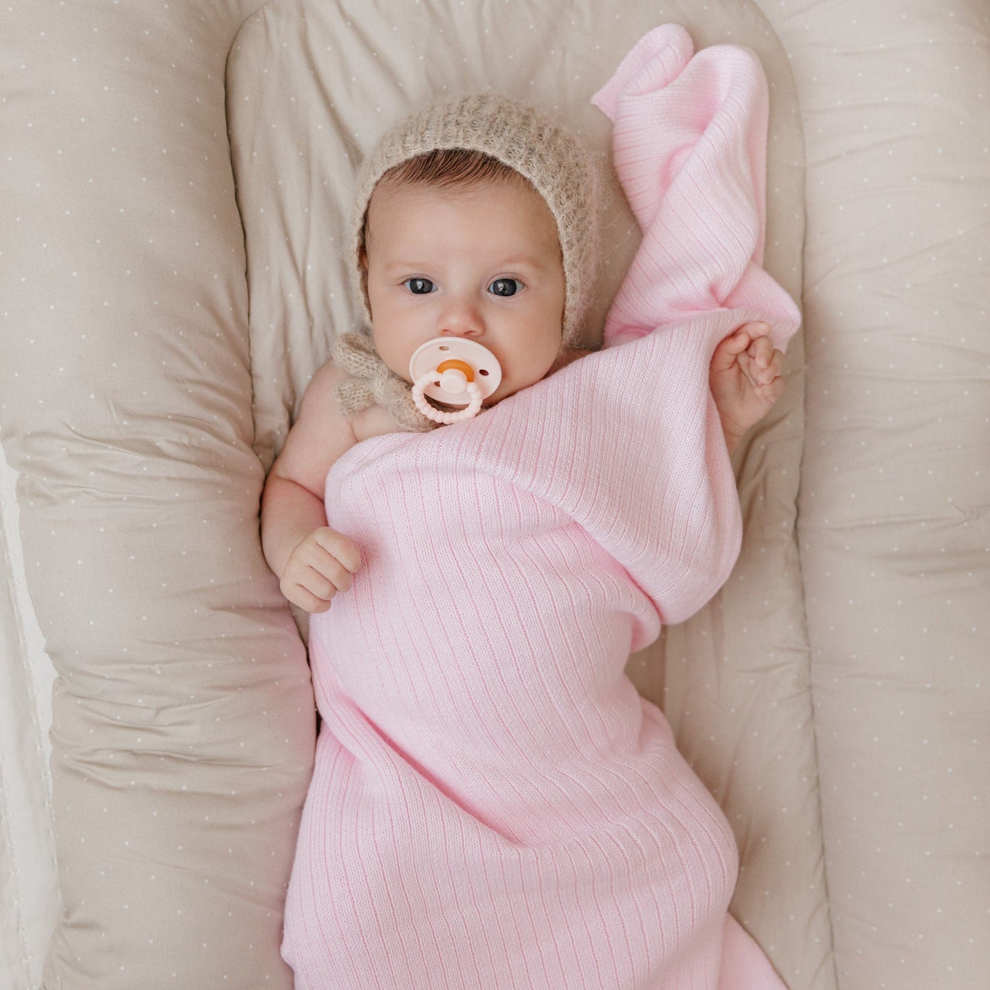 Baby wrapped in a pink blanket with a pacifier, lying on a beige surface.