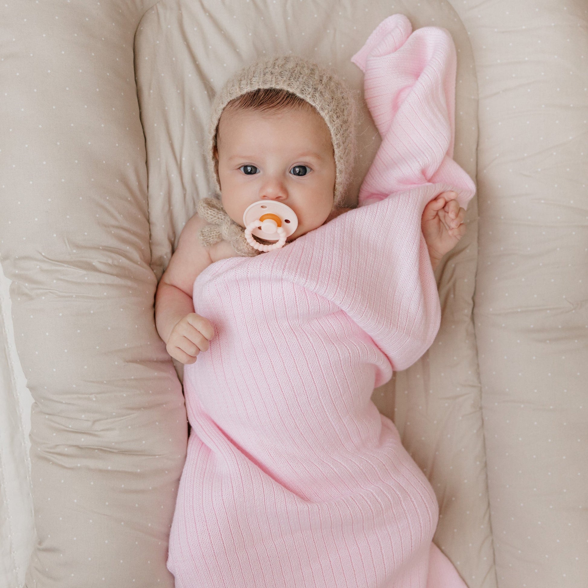 Baby wrapped in a pink blanket with a pacifier, lying on a beige surface.