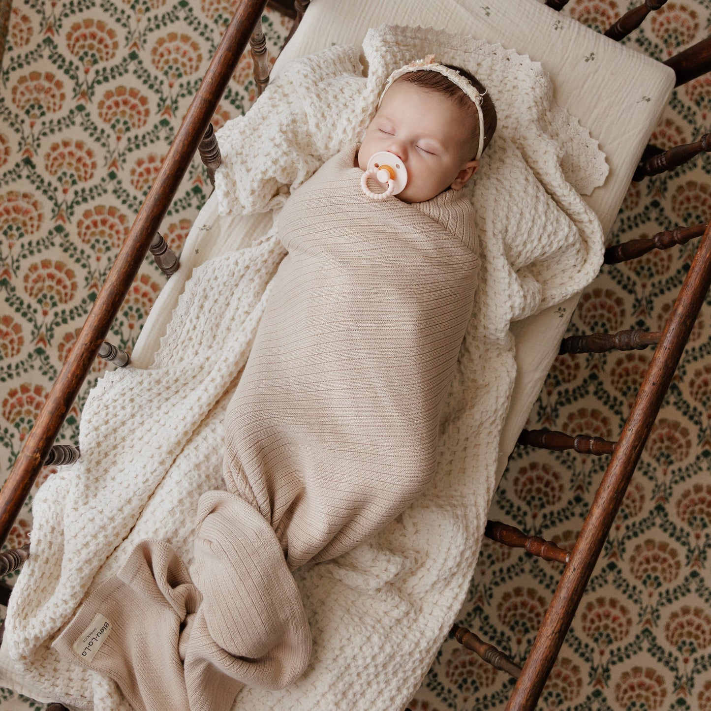 Newborn baby swaddled in beige blanket with pacifier, lying in a wooden crib.