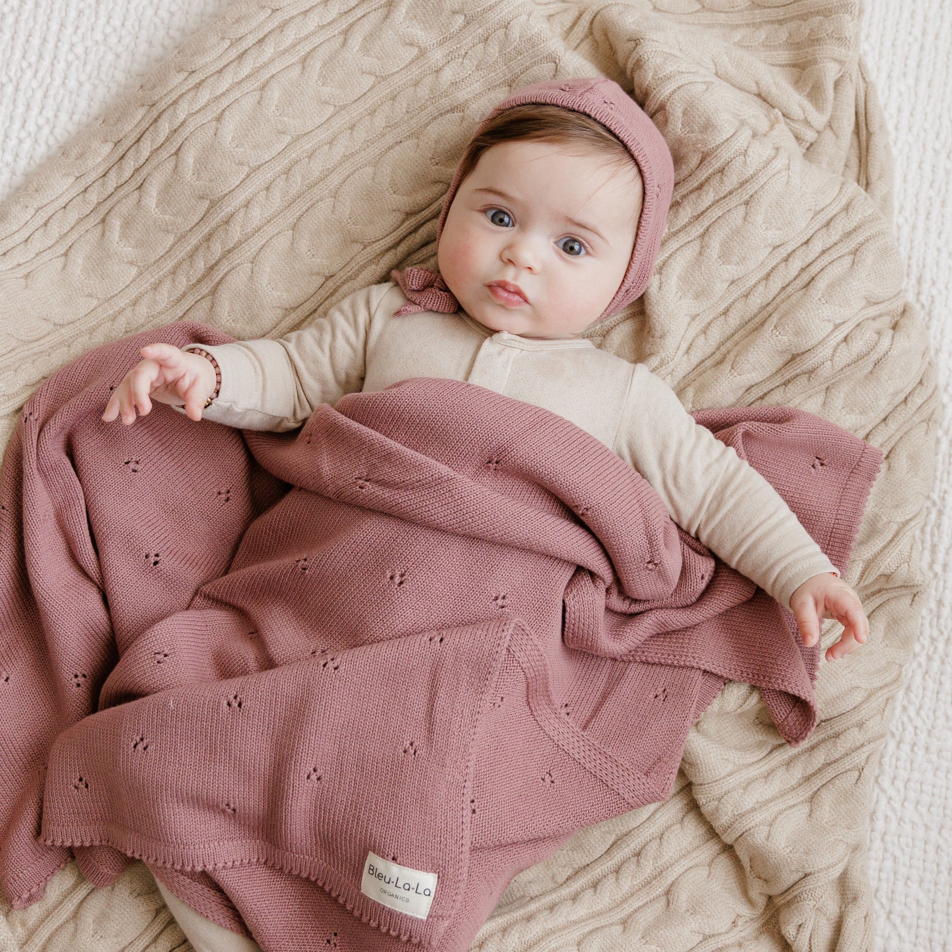 Baby lying on a textured surface with a pink blanket and headband, wearing a beige outfit.