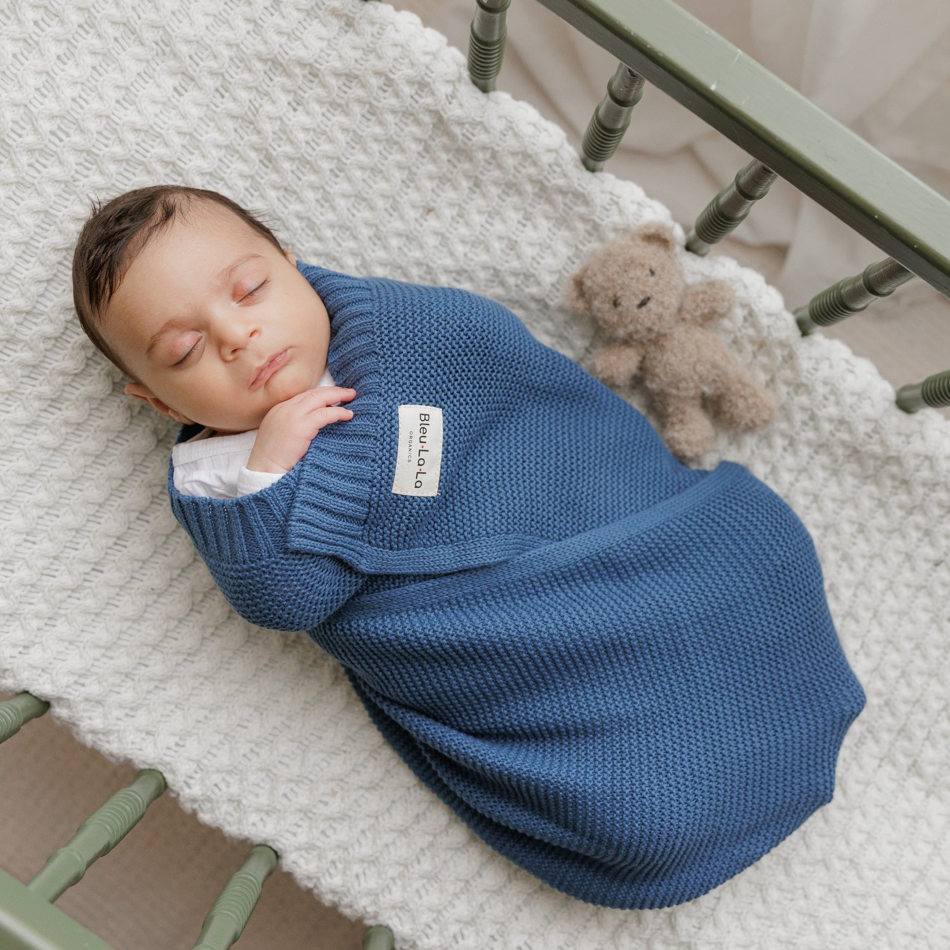 Newborn baby swaddled in a blue Blue-La-Lablanket with a teddy bear on a crib