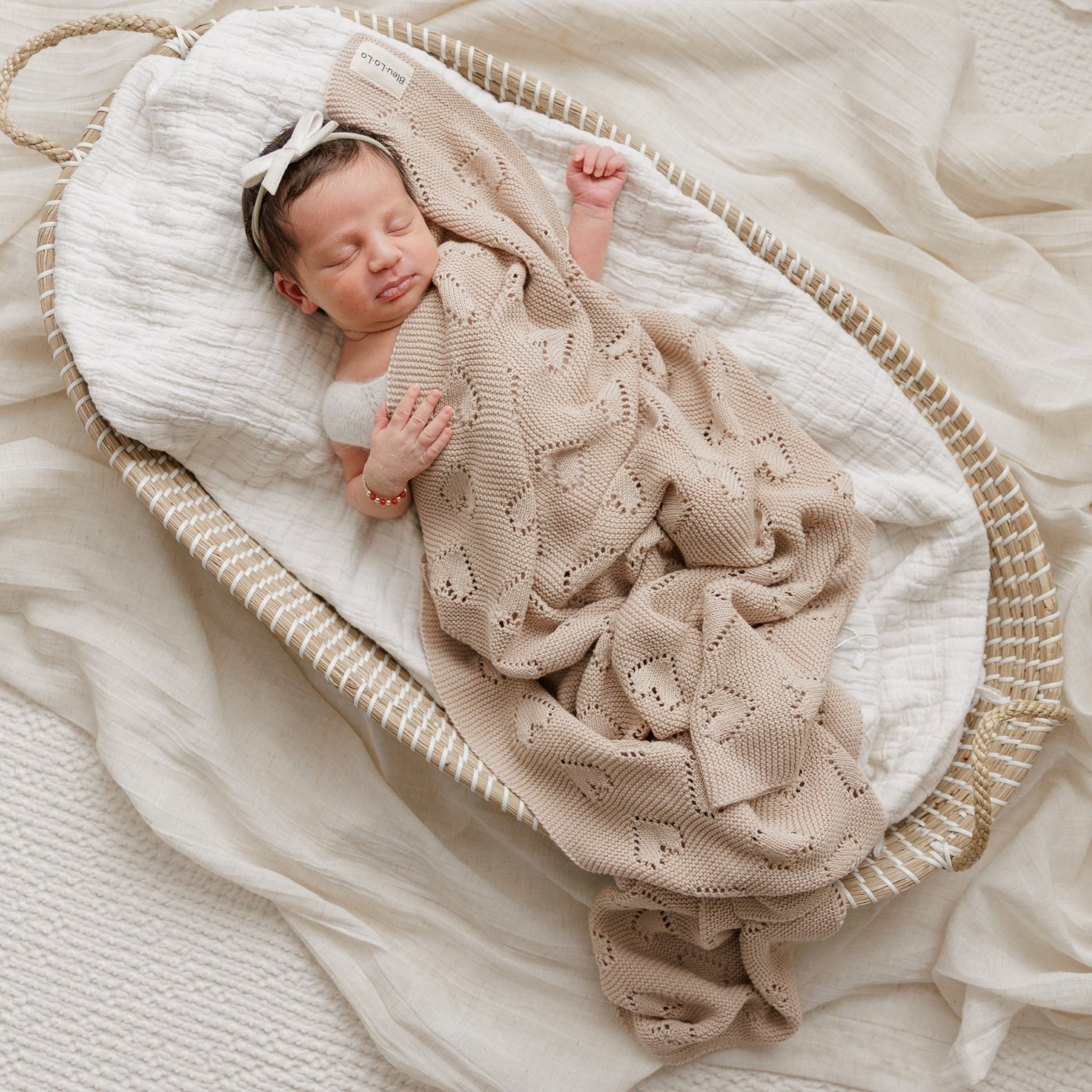 Newborn baby wrapped in a beige Bleu-la-la knitted blanket in a white crib.