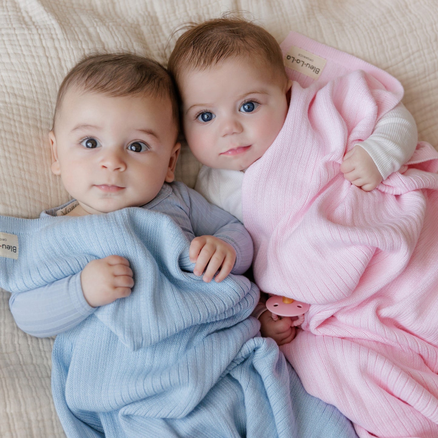 Two babies wrapped in blue and pink blankets on a beige blanket