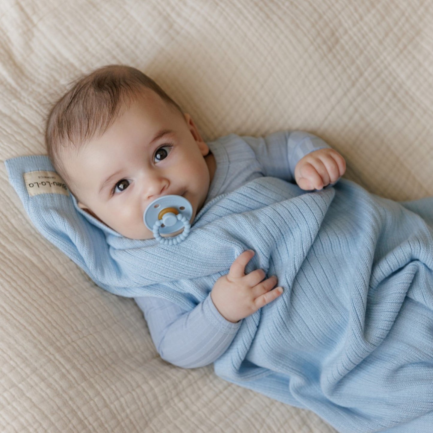Baby wrapped in a blue blanket with a pacifier, lying on a beige textured surface.
