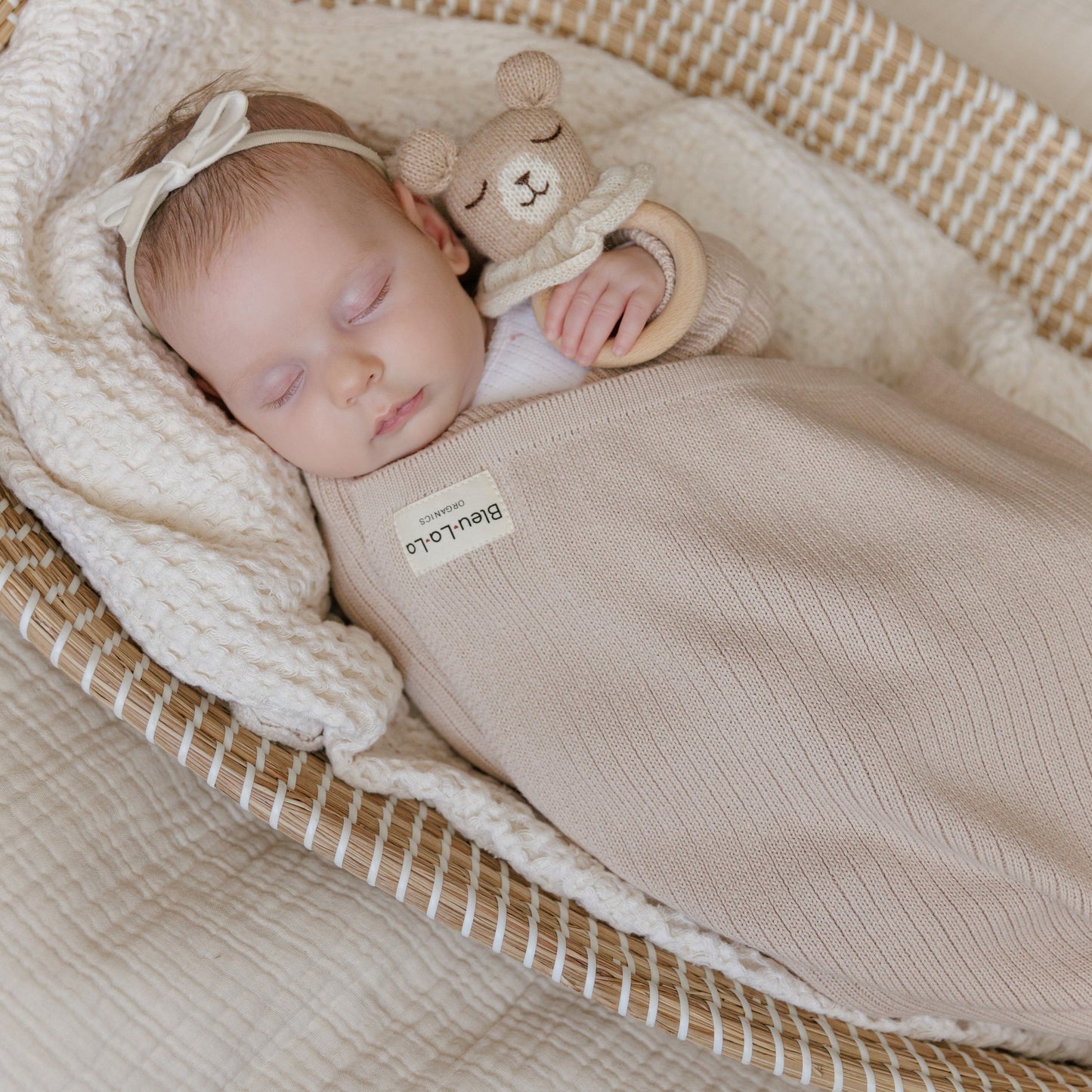Newborn baby sleeping in a crib with a beige swaddle and teddy bear, wearing a headband.