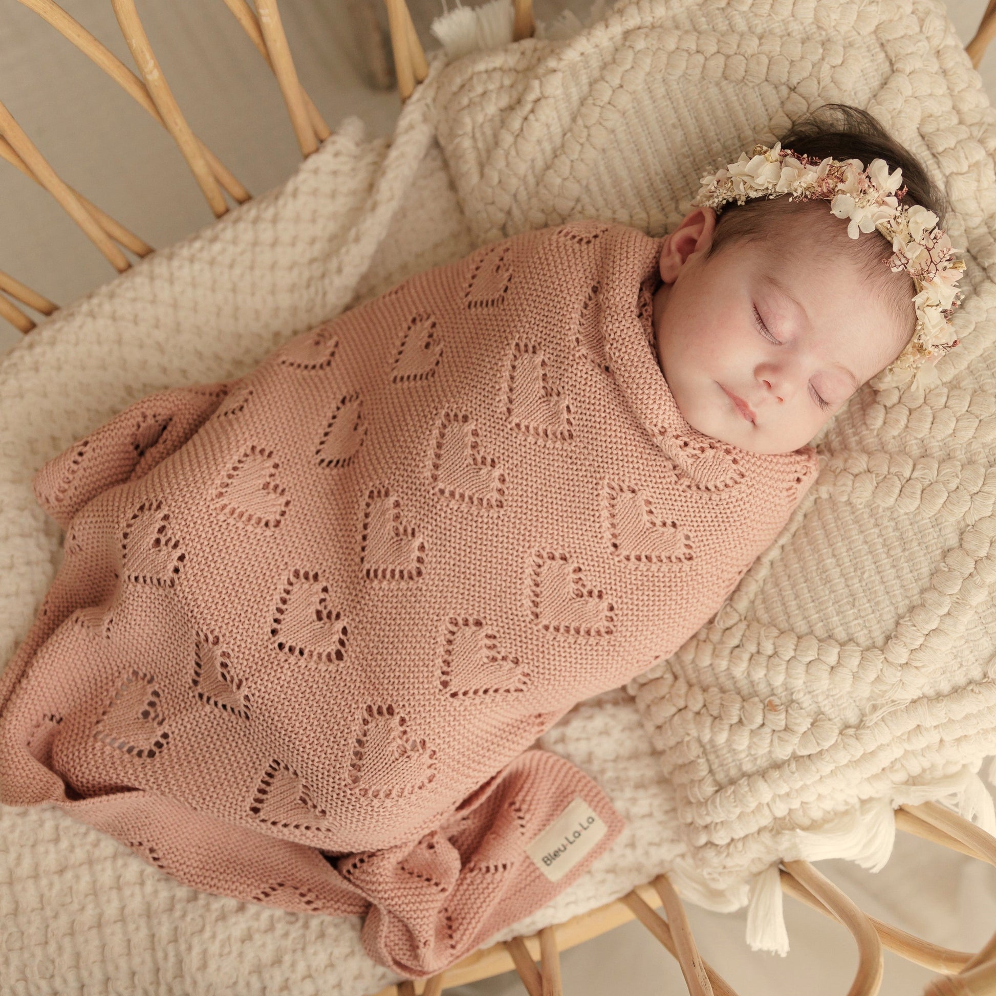 Newborn baby wrapped in a pink Bleu-la-la knitted blanket with heart patterns, lying on a textured surface.