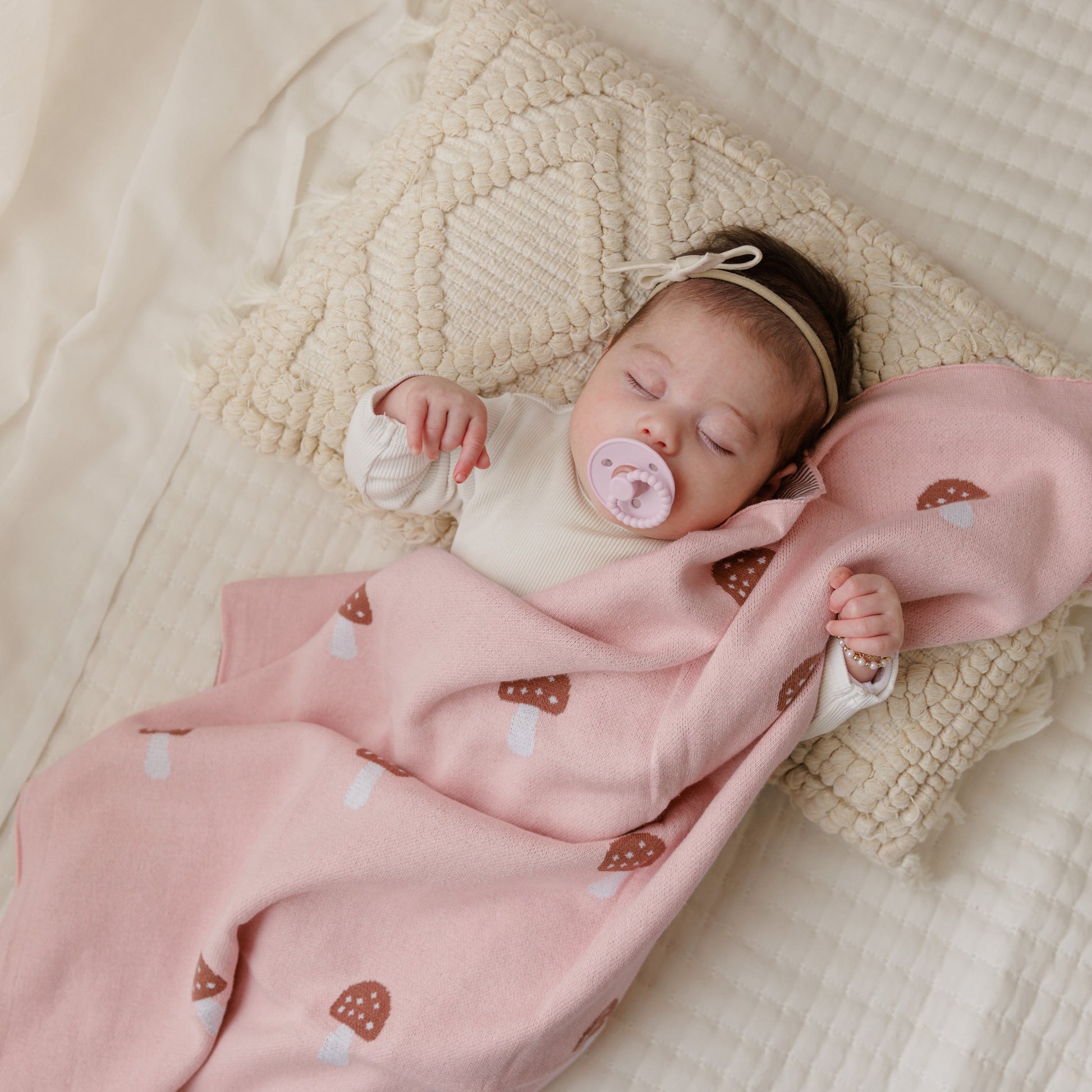 Baby sleeping under a pink blanket with mushroom patterns on a beige textured surface