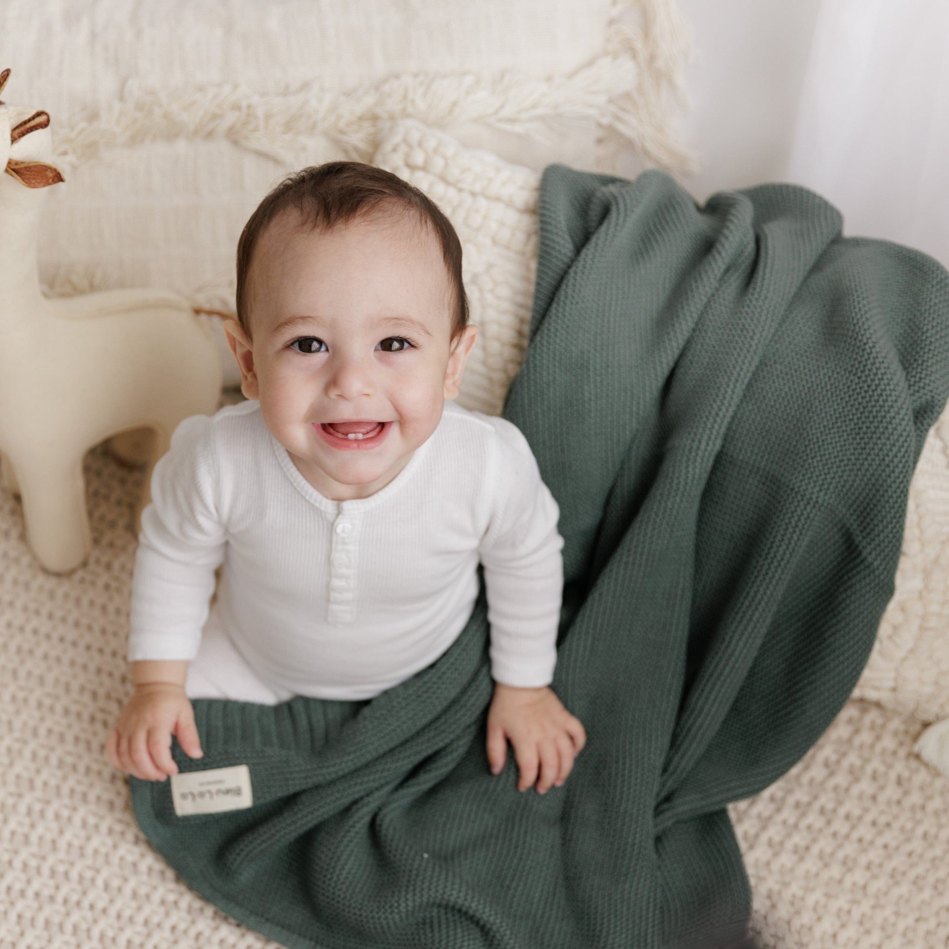 Baby in a white outfit lying on a green Blue-La-La blanket with a neutral background