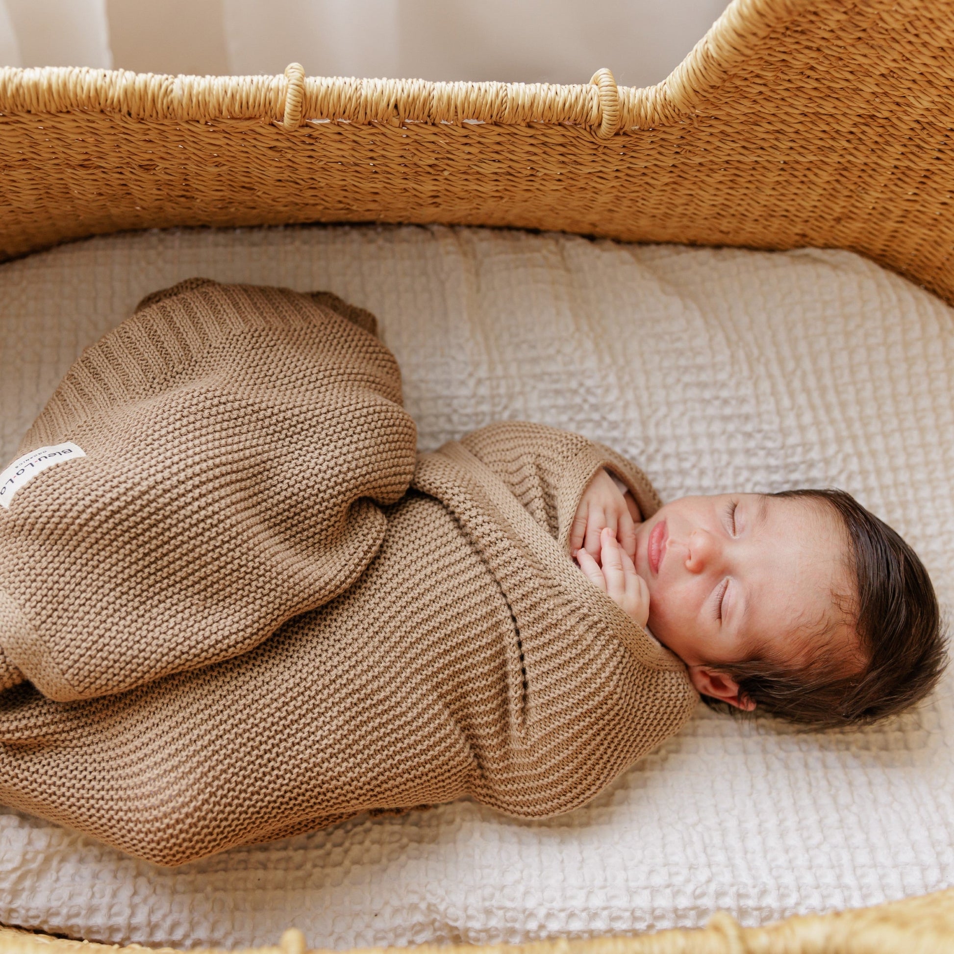 Newborn baby wrapped in a brown Blue-La-La blanket lying in a woven basket.