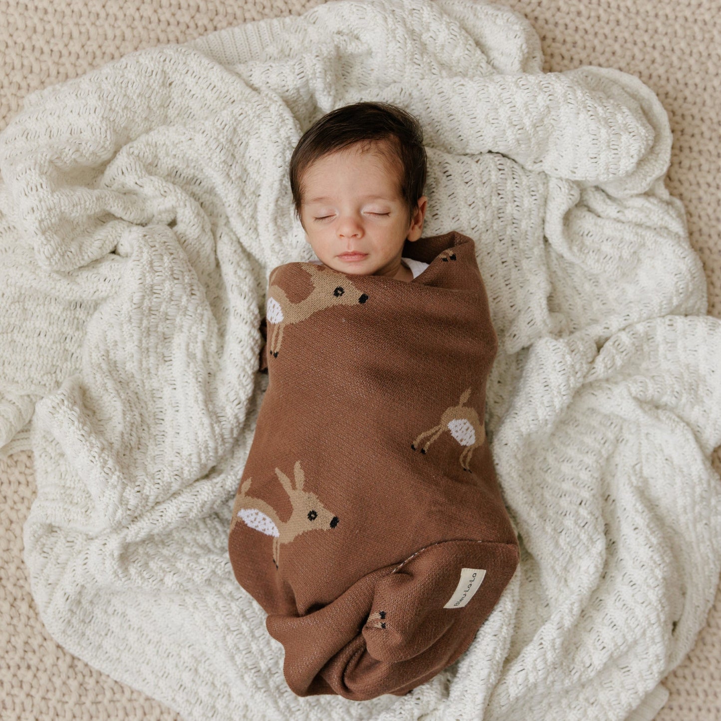 Newborn baby swaddled in a brown blanket with deer patterns on a soft beige blanket.