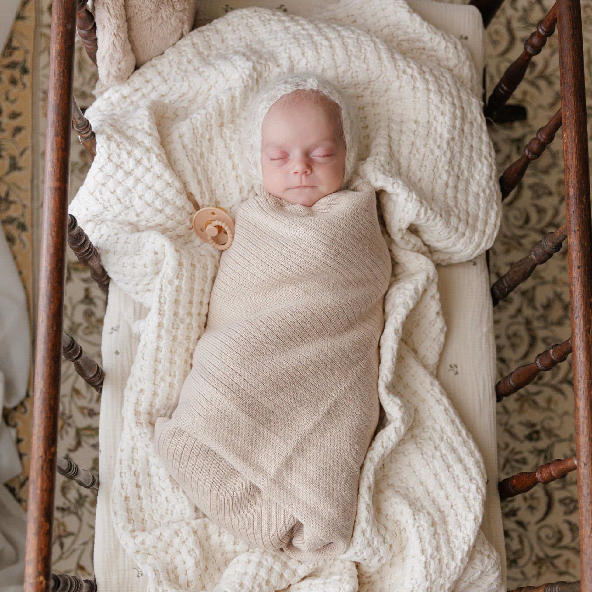 Newborn baby swaddled in a beige blanket in a wooden crib with a textured wall in the background