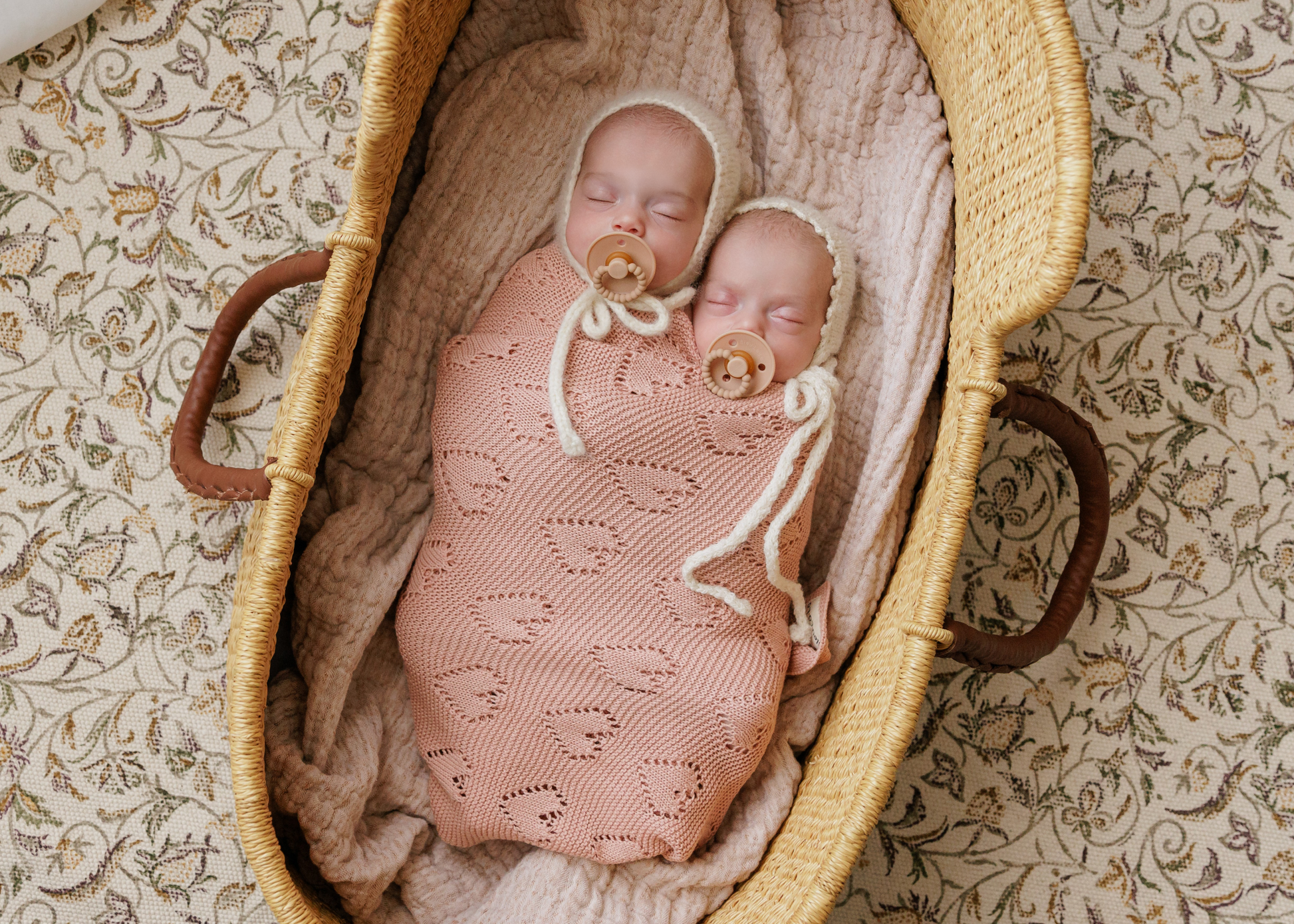 Two babies wrapped in pink  Bleu-la-la blankets with pacifiers in a wicker basket on a patterned surface.