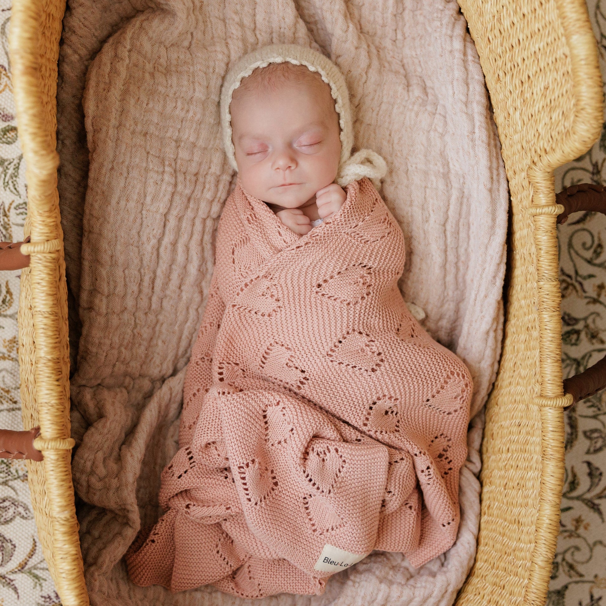 Newborn baby wrapped in a pink Bleu-la-la knitted blanket and bonnet, lying in a moses basket.