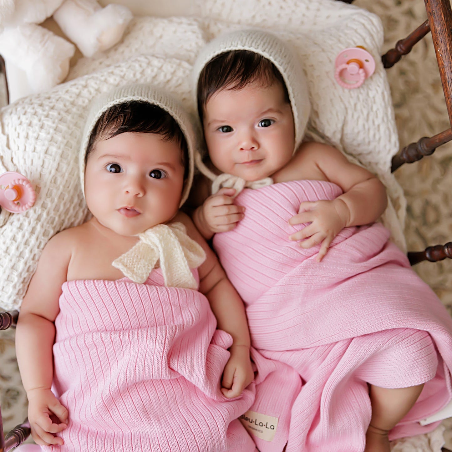 Two babies wrapped in pink blankets with knitted hats, sitting on a wooden chair.