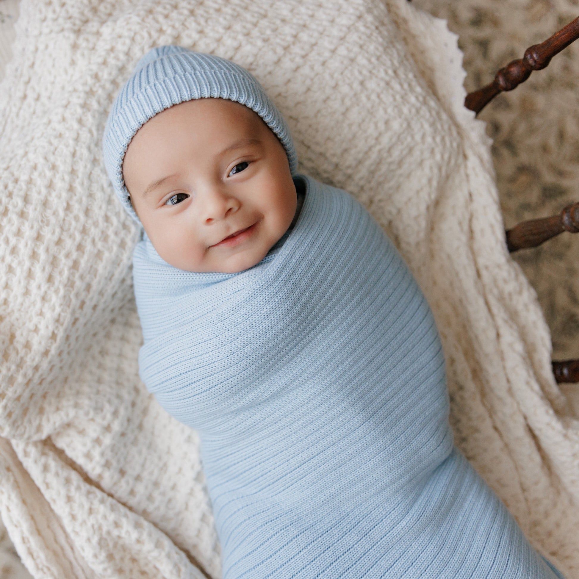 Baby wrapped in a light blue swaddle and hat, lying on a textured beige blanket.