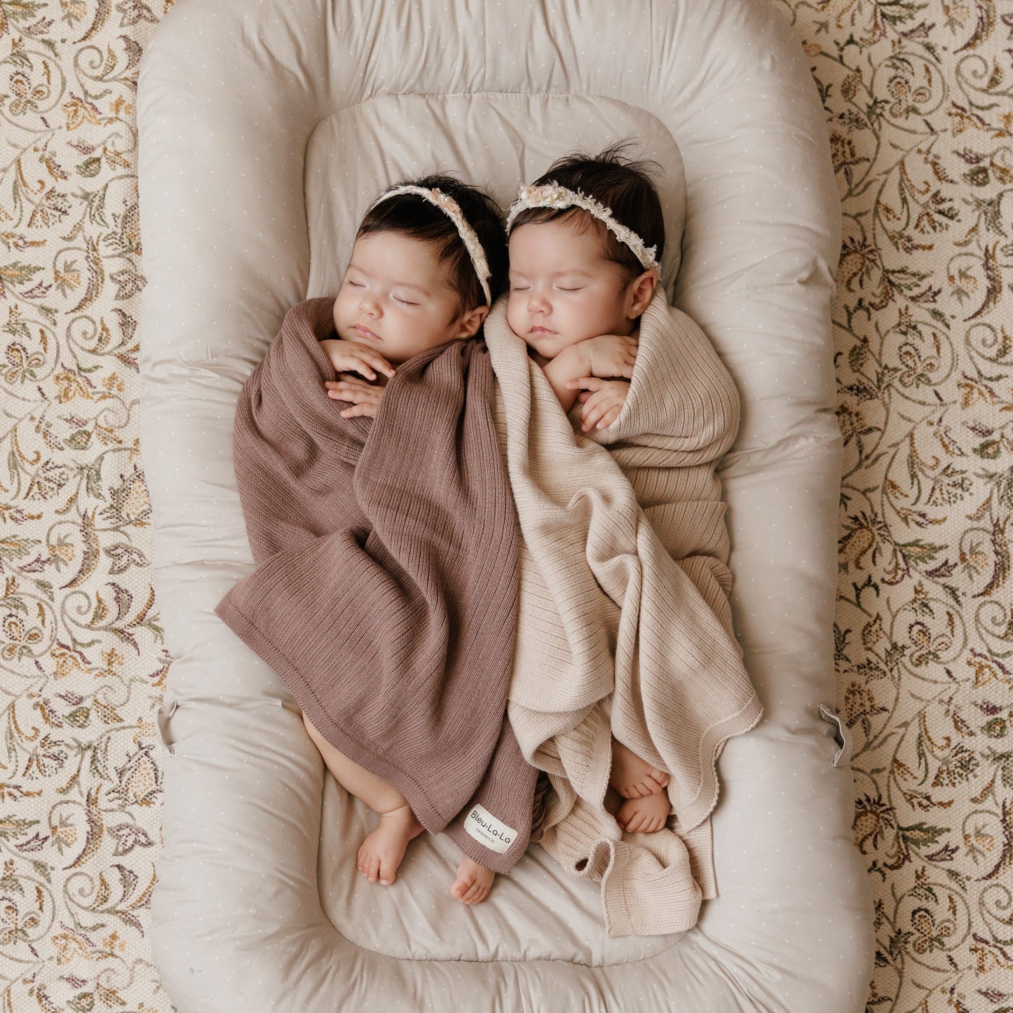 Two babies wrapped in blankets lying on a cushion with a patterned background