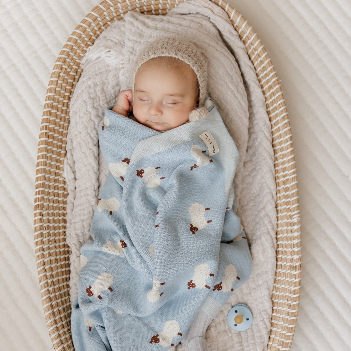 Newborn baby wrapped in a blue blanket with sheep patterns, sleeping in a wicker crib.