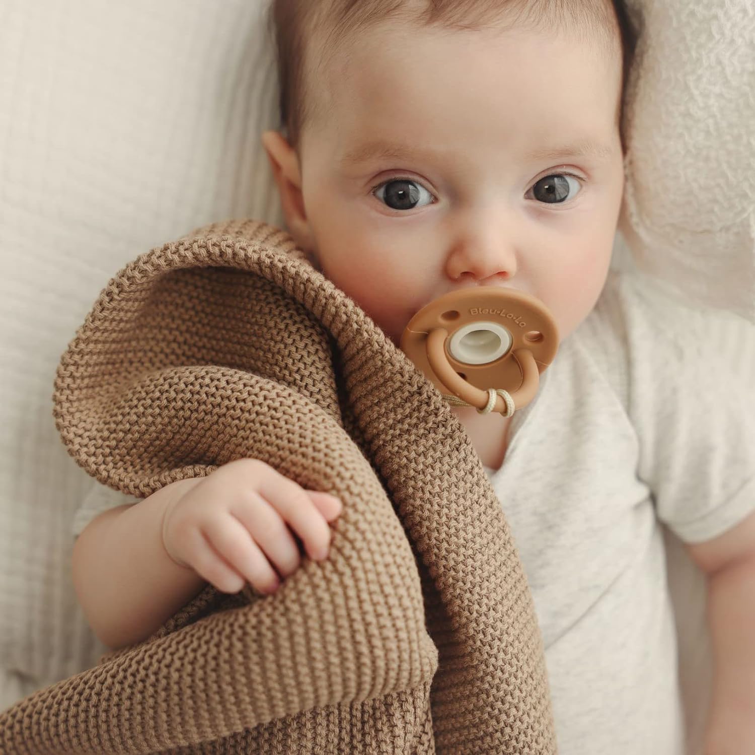 Baby wrapped in a brown blanket Blue-La-La with a wooden pacifier