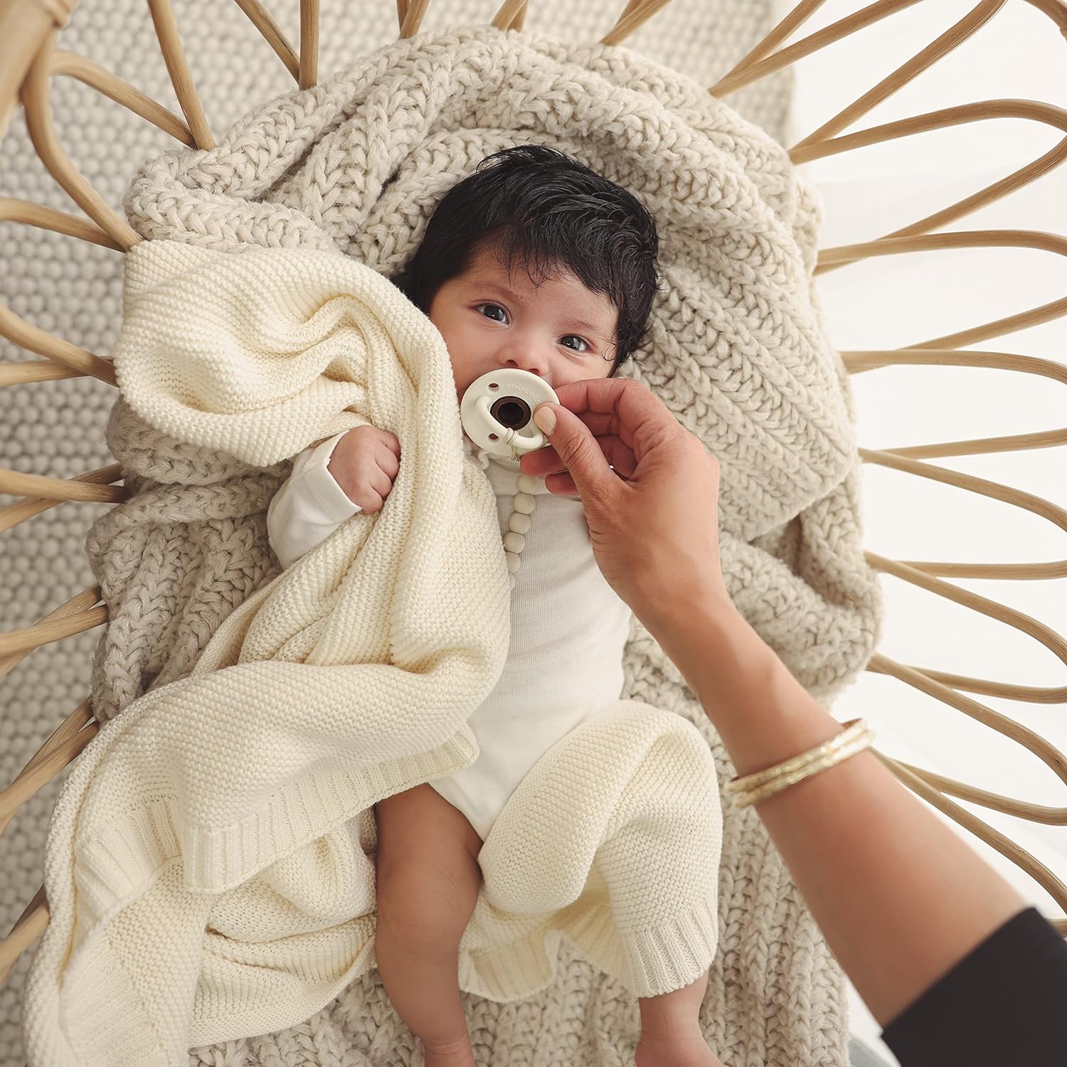 Baby wrapped in a white Blue-La-La blanket with a pacifier, sitting in a wicker chair.