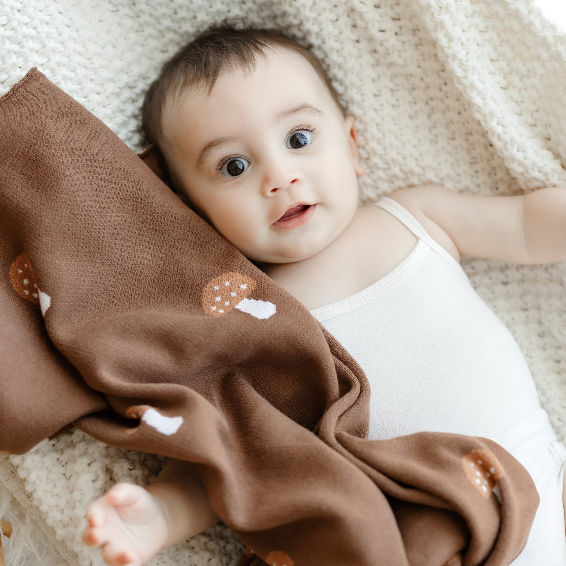 Baby lying on a soft surface with a brown blanket featuring mushroom patterns.