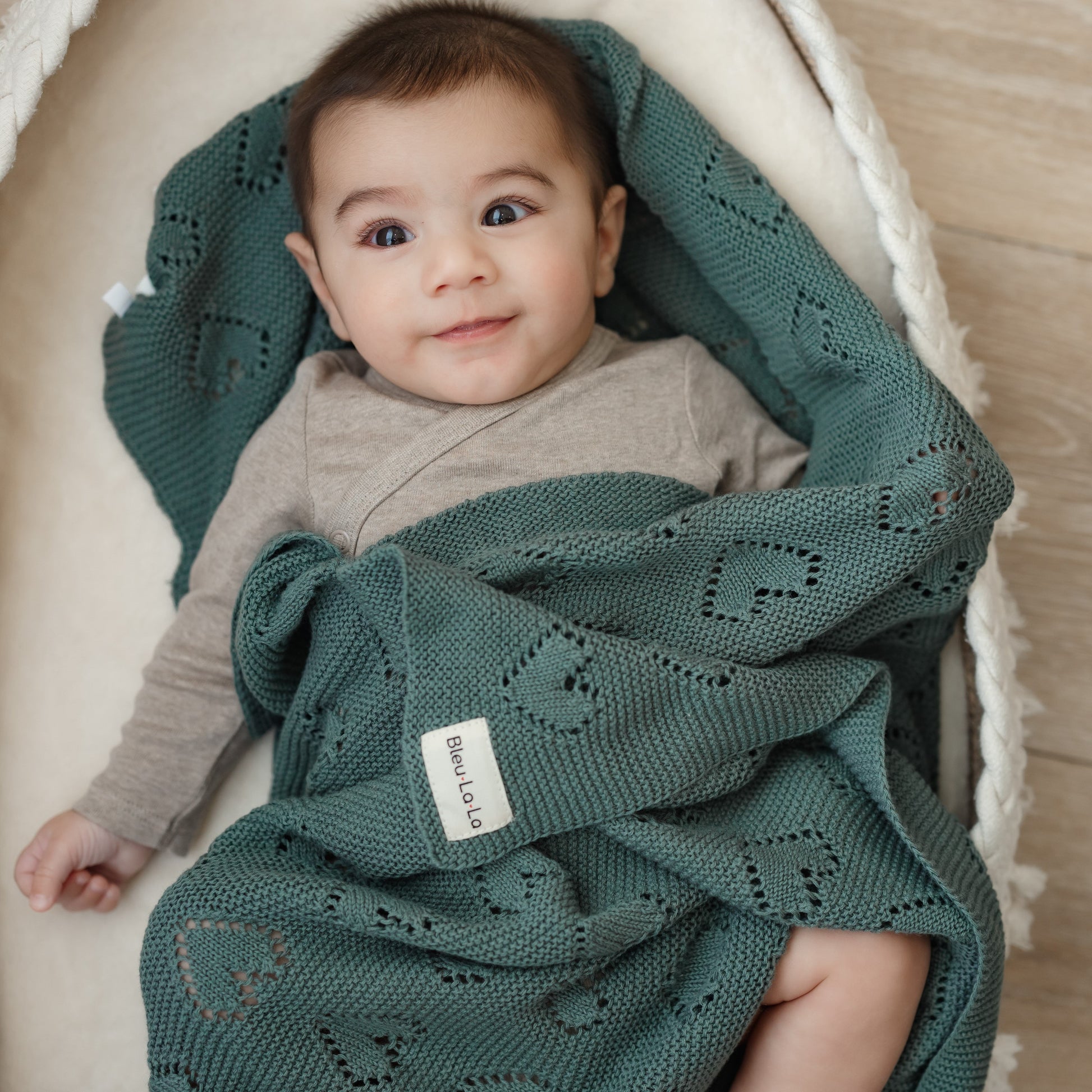 Baby wrapped in a green knitted Bleu-la-la blanket with a visible brand label, sitting in a white crib.