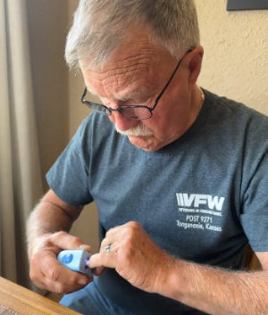 A senior veteran with glasses and a VFW t-shirt on is using Lil Nipper Adult clipper, featuring a quiet motor for a relaxing and discreet nail trimming experience. He's concentrating and inserting his fingernail into the safety slot. He looks strong.